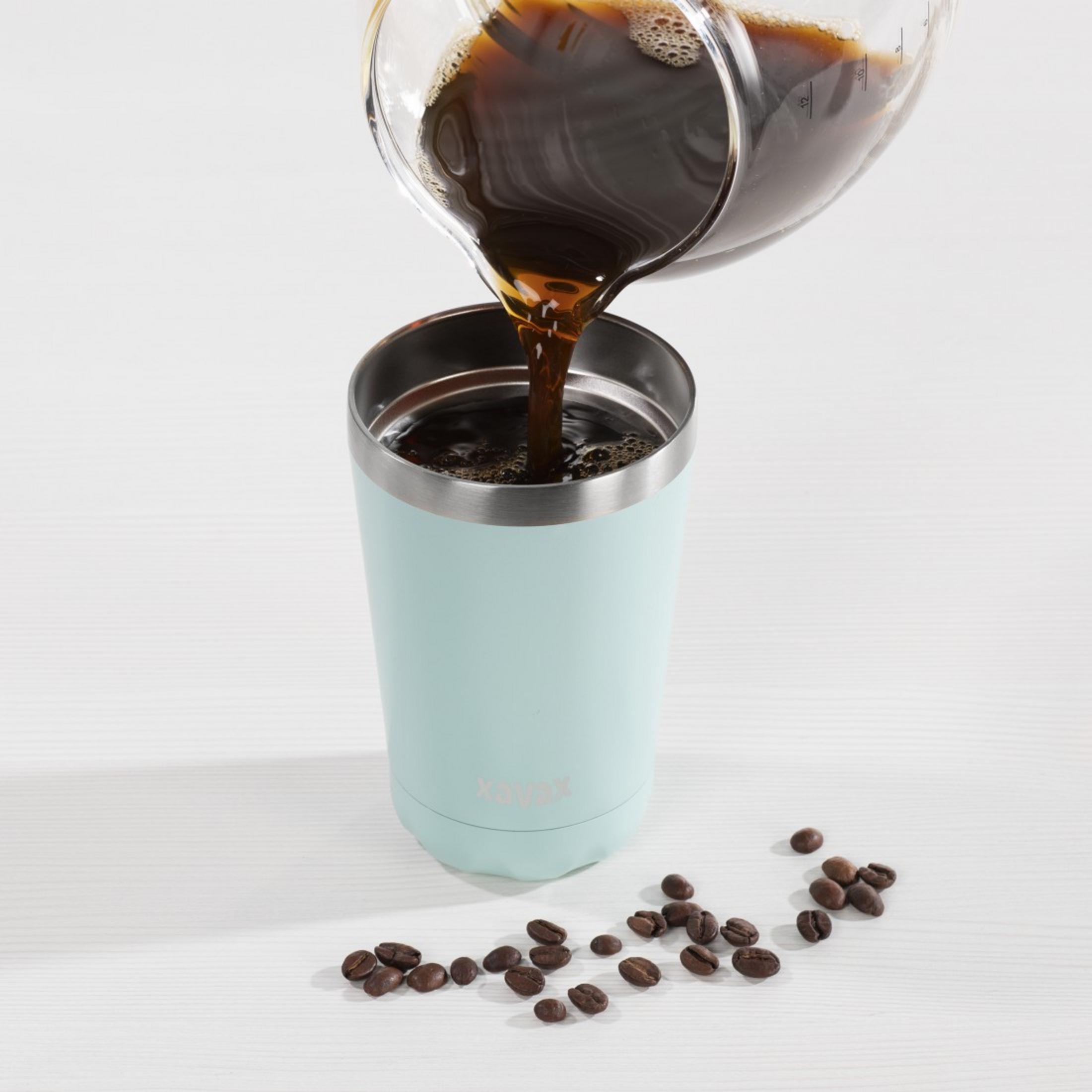 Kaffee wird aus einem Glas in einen hellblauen Becher gegossen. Kaffeebohnen liegen daneben.