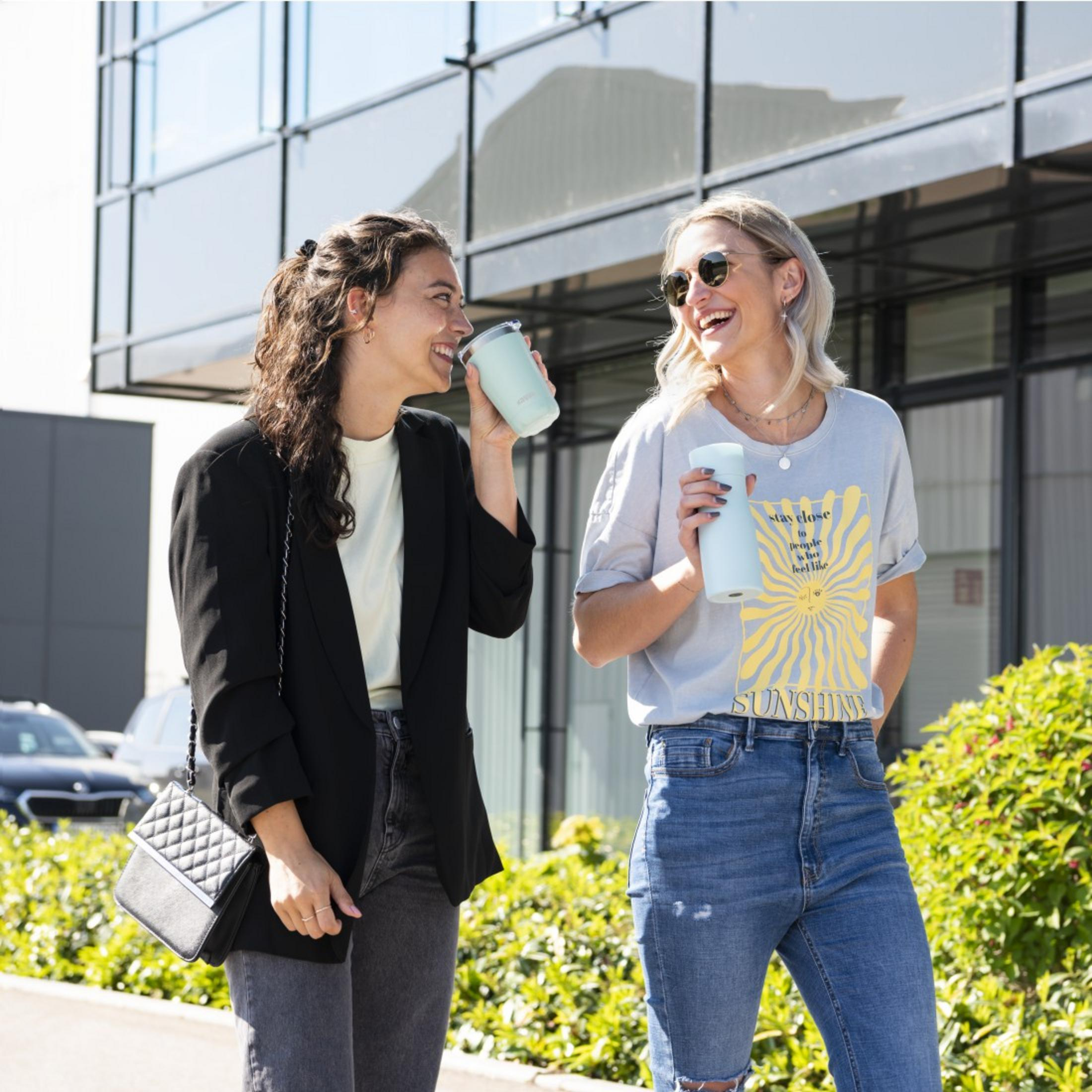 Zwei Frauen trinken aus hellblauen Bechern vor einem modernen Gebäude.