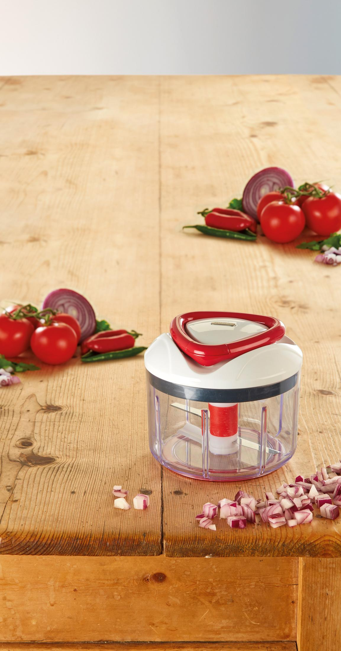 Un tritatutto da cucina trasparente con manico rosso è su un tavolo di legno vicino a pomodori e cipolle.
