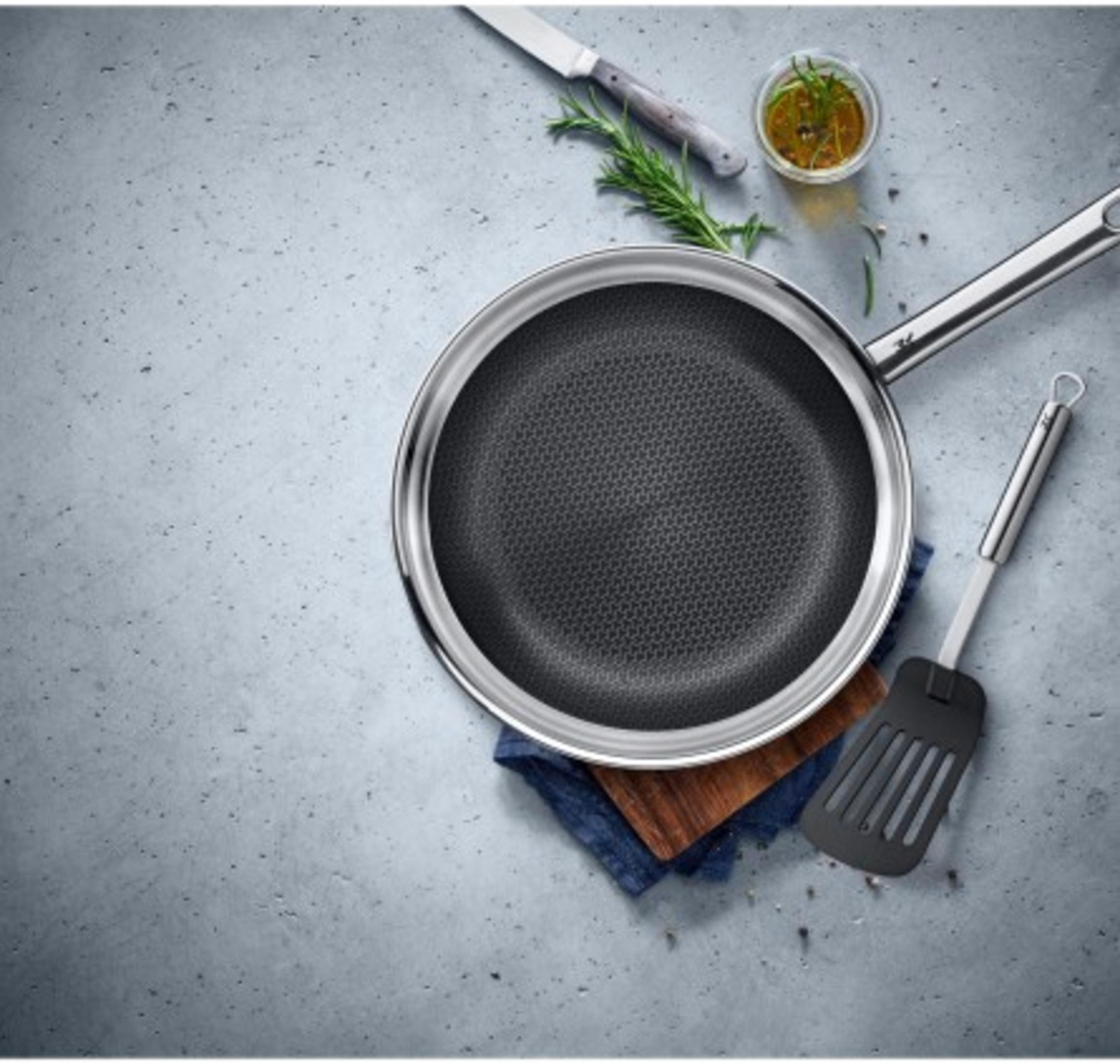 Patelnia z wzorem plastra miodu, nóż, rozmaryn, olej i łopatka na szarej powierzchni.