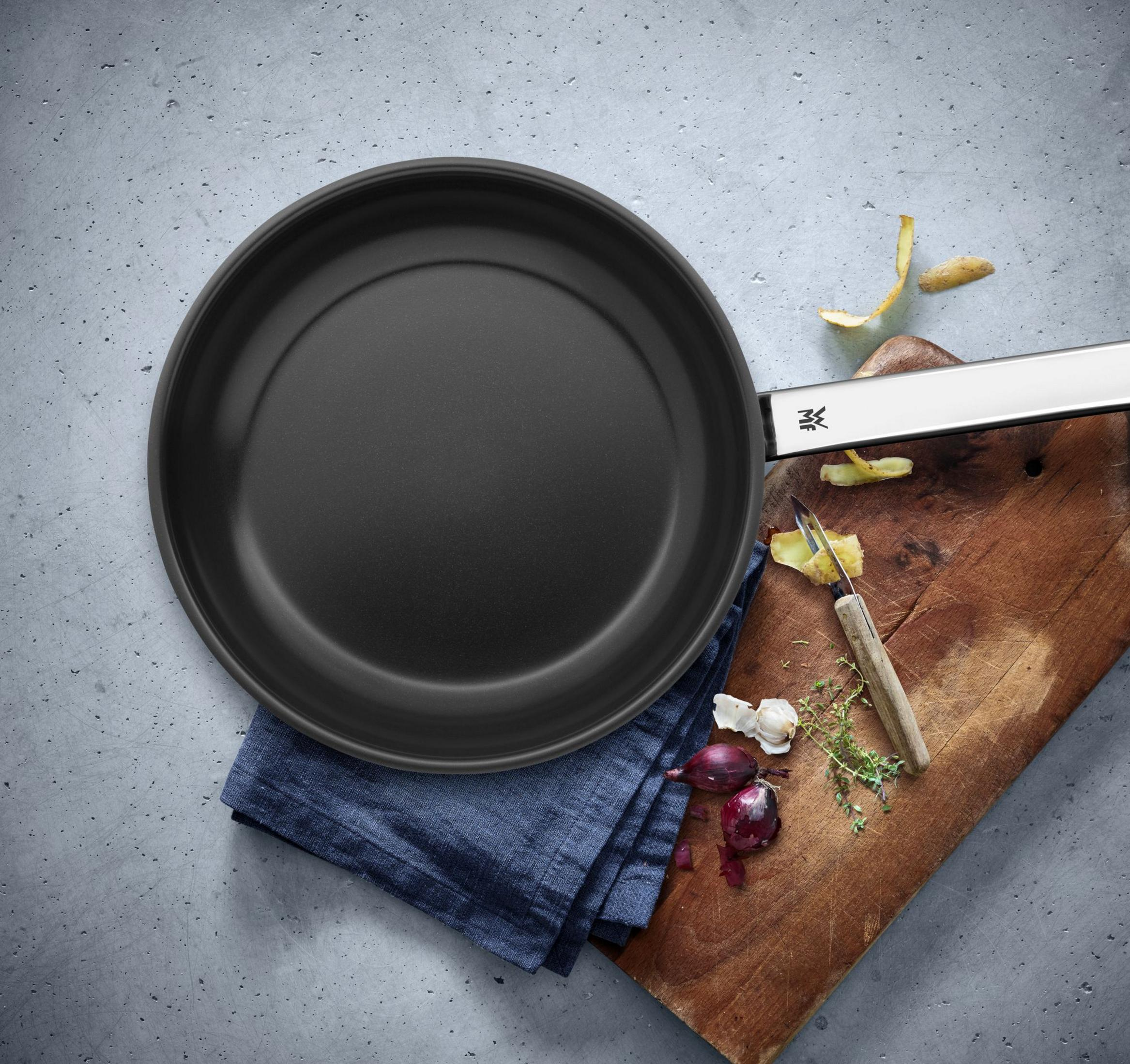 Schwarze Pfanne auf blauem Tuch, neben einem Holzbrett mit Zwiebeln, Knoblauch, Kartoffelschalen und einem Sparschäler.
