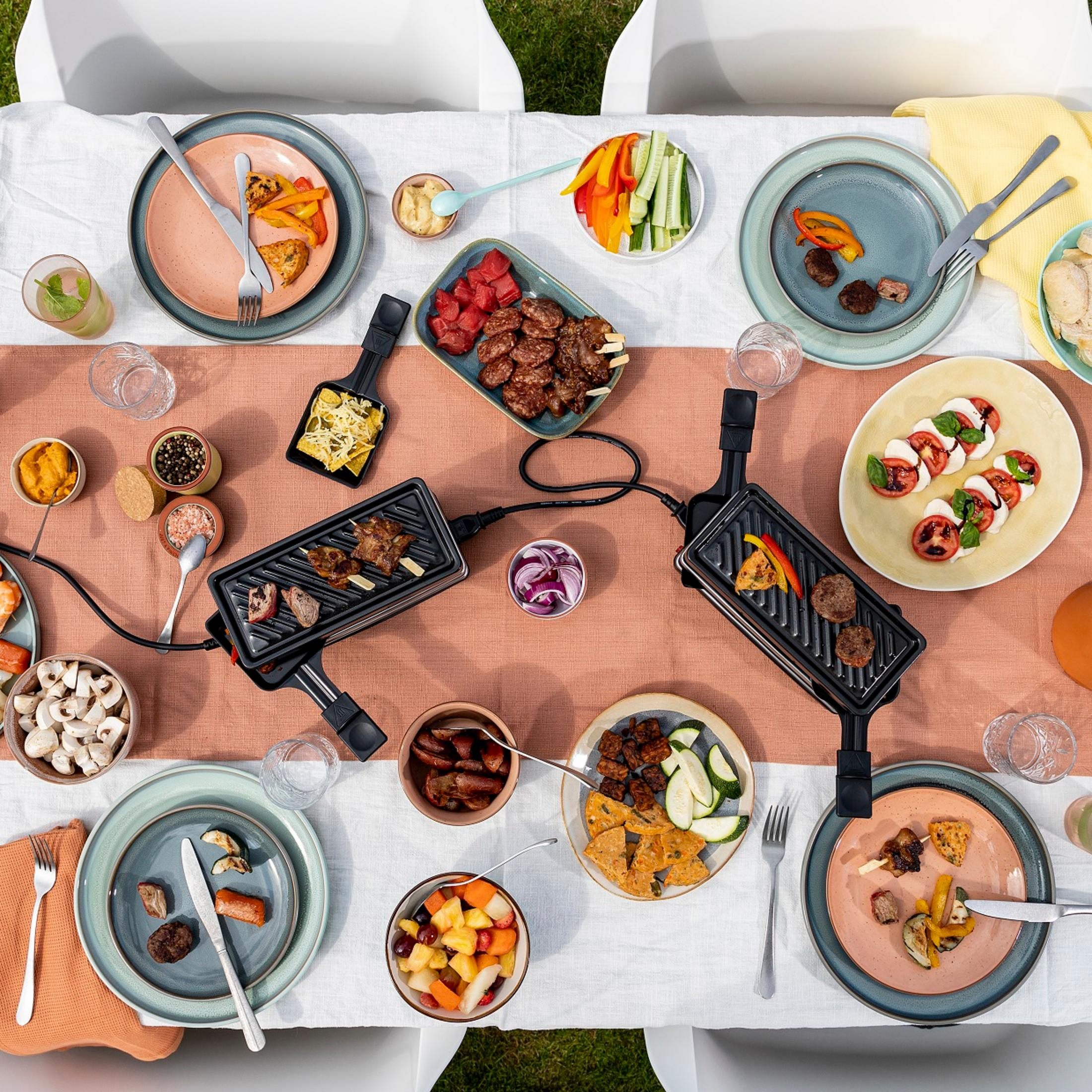 Tavola apparecchiata con griglie per raclette, carne, verdure, insalate e frutta.