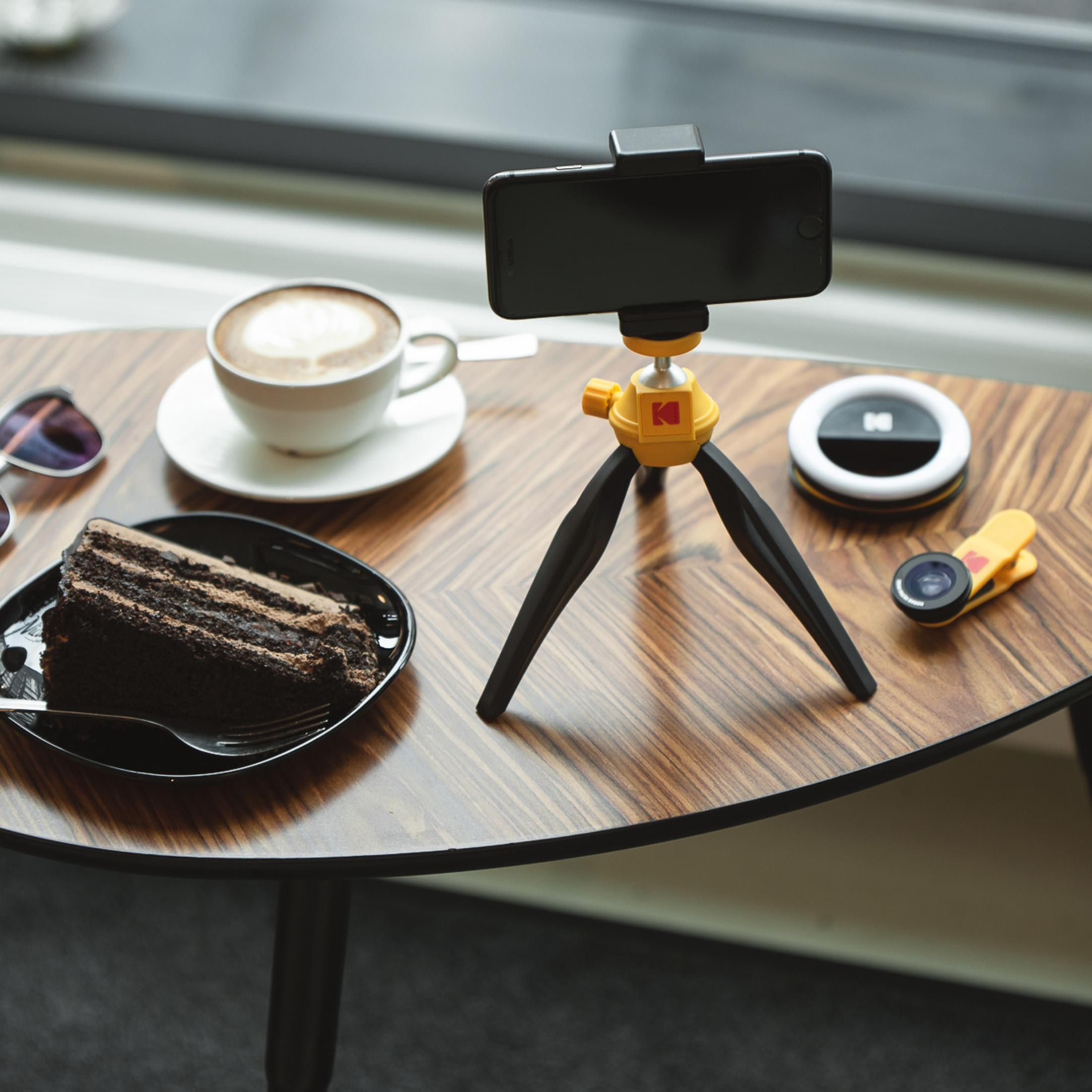 Una rebanada de pastel de chocolate, café y un trípode para smartphone en una mesa de madera.