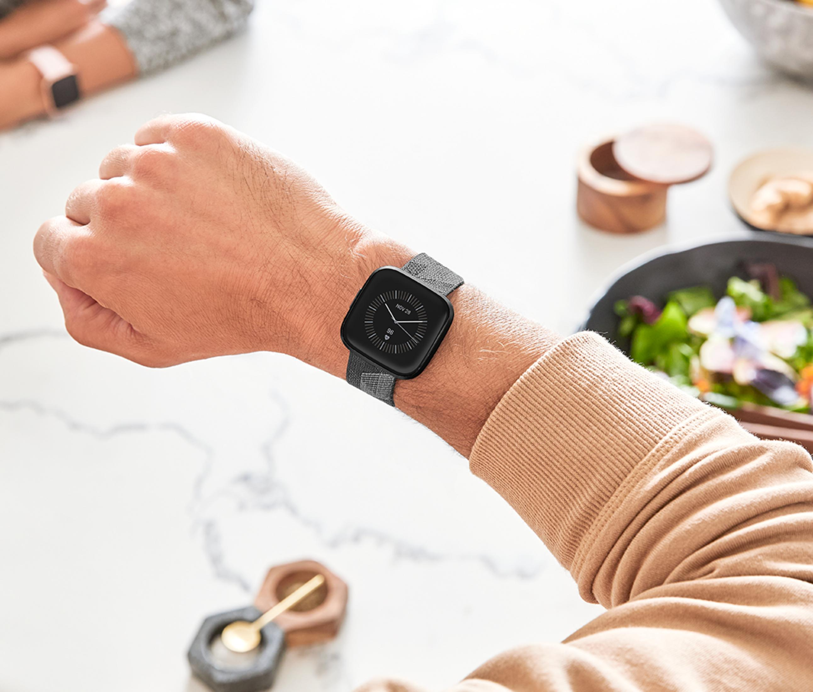 Una mano lleva un reloj inteligente con correa gris. Ensalada al fondo.