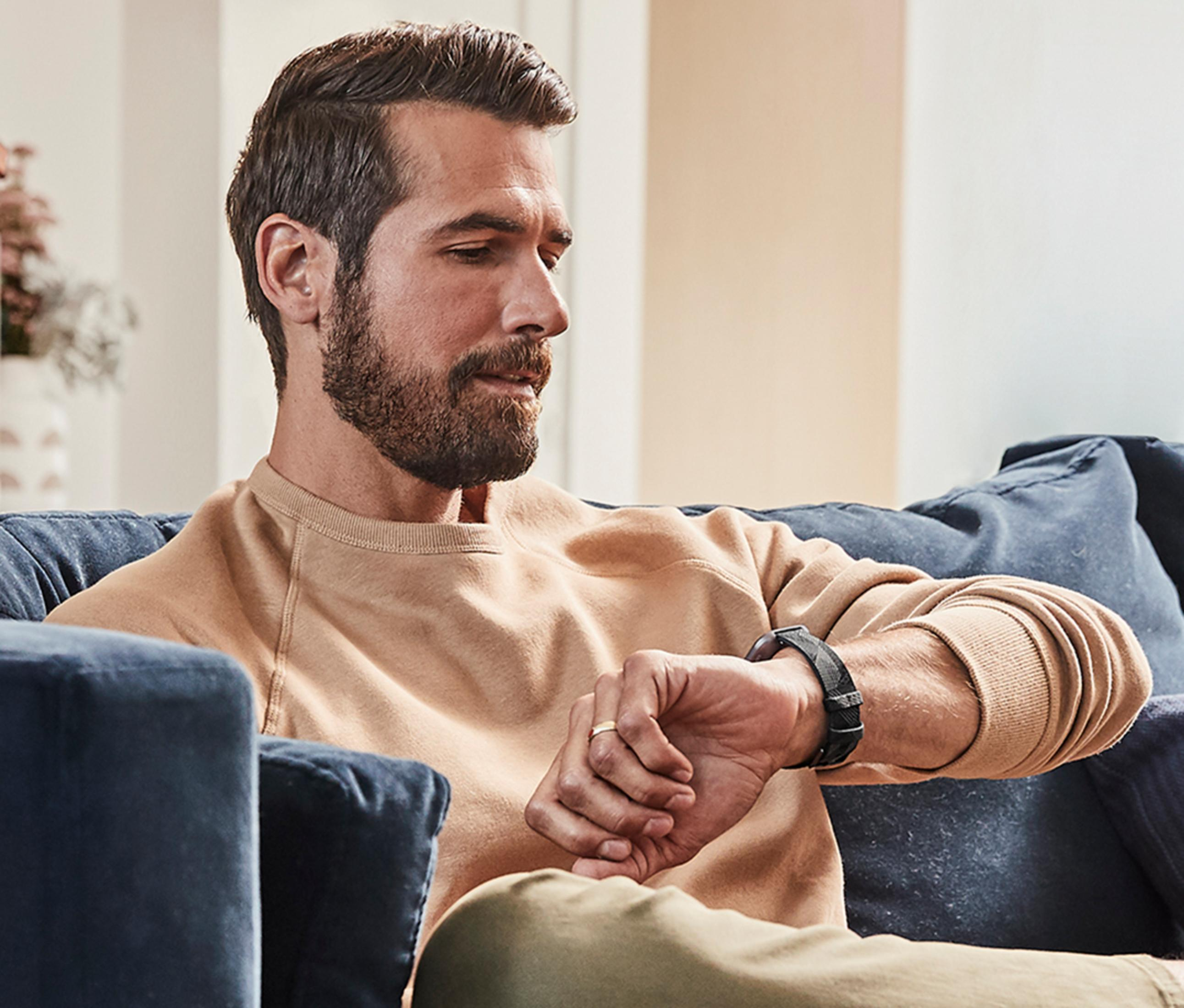 Un hombre con barba se sienta en un sofá mirando su reloj inteligente.