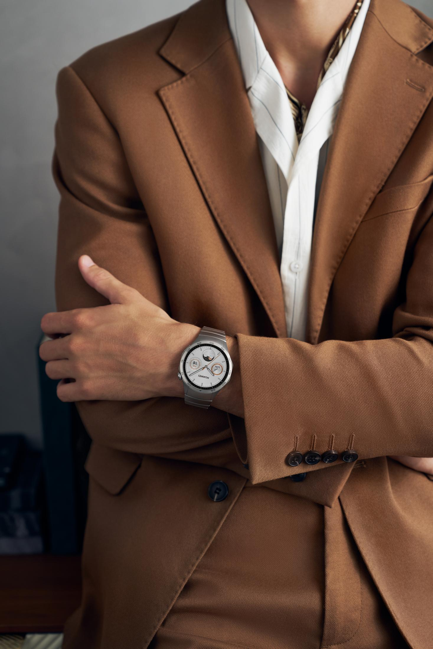 Mężczyzna w brązowym garniturze nosi srebrny smartwatch na lewym nadgarstku, ręce skrzyżowane.