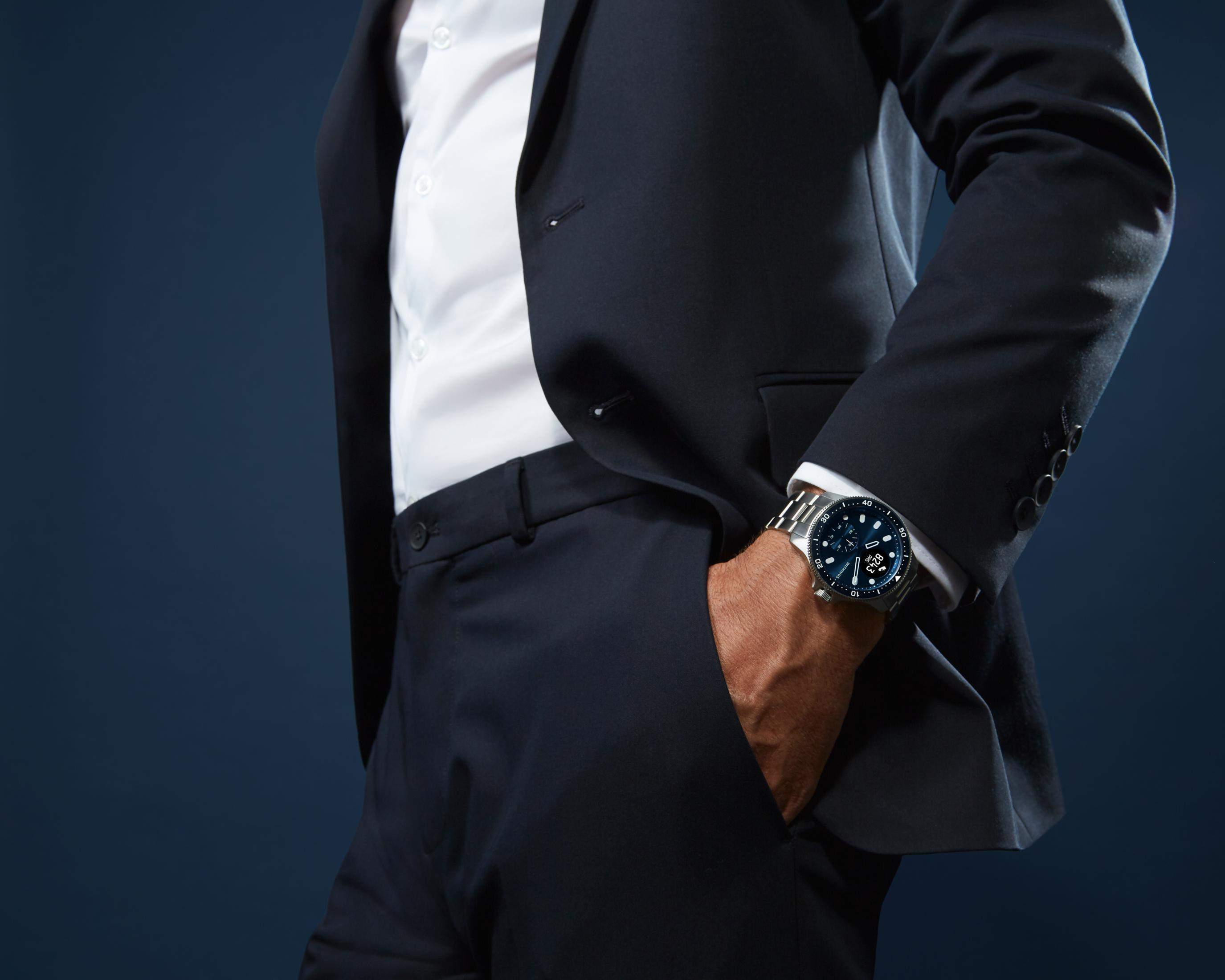 Mann im dunklen Anzug mit weißem Hemd und silberner Uhr. Hand in Hosentasche.