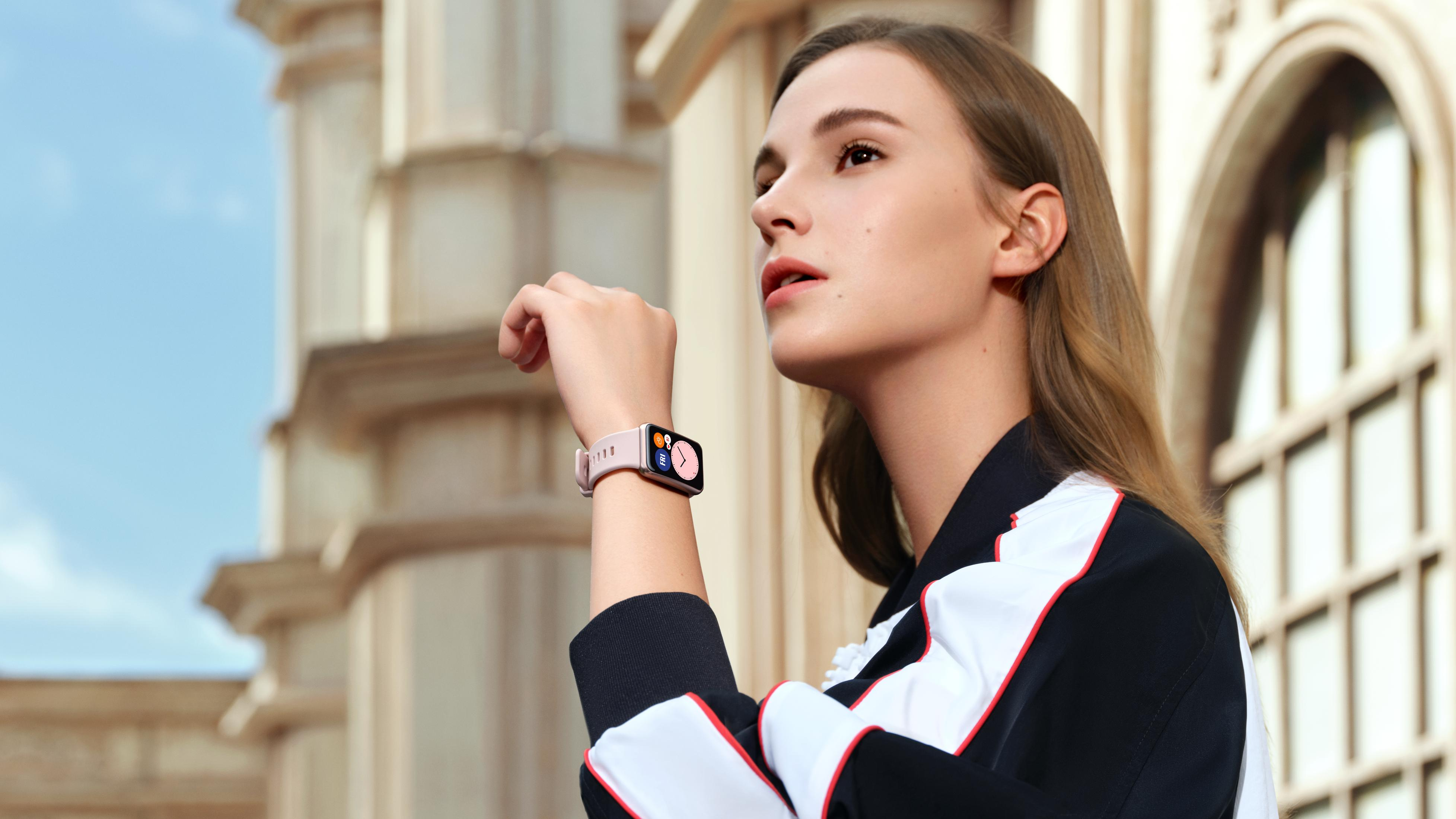 Eine junge Frau mit einem rosa Smartwatch vor einer hellen Fassade.