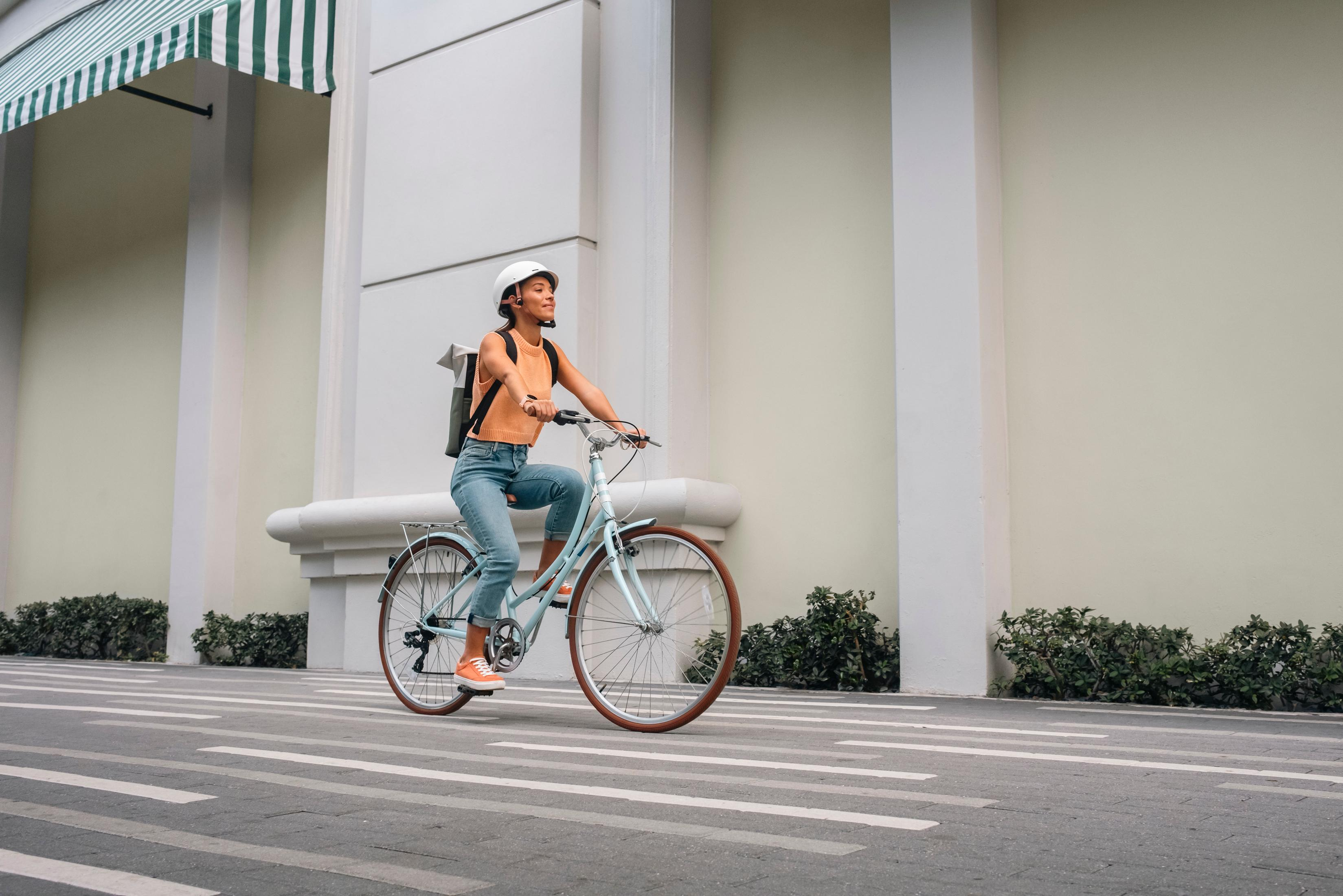 Una mujer con casco monta en bicicleta por una calle con rayas. Viste jeans y un top naranja.