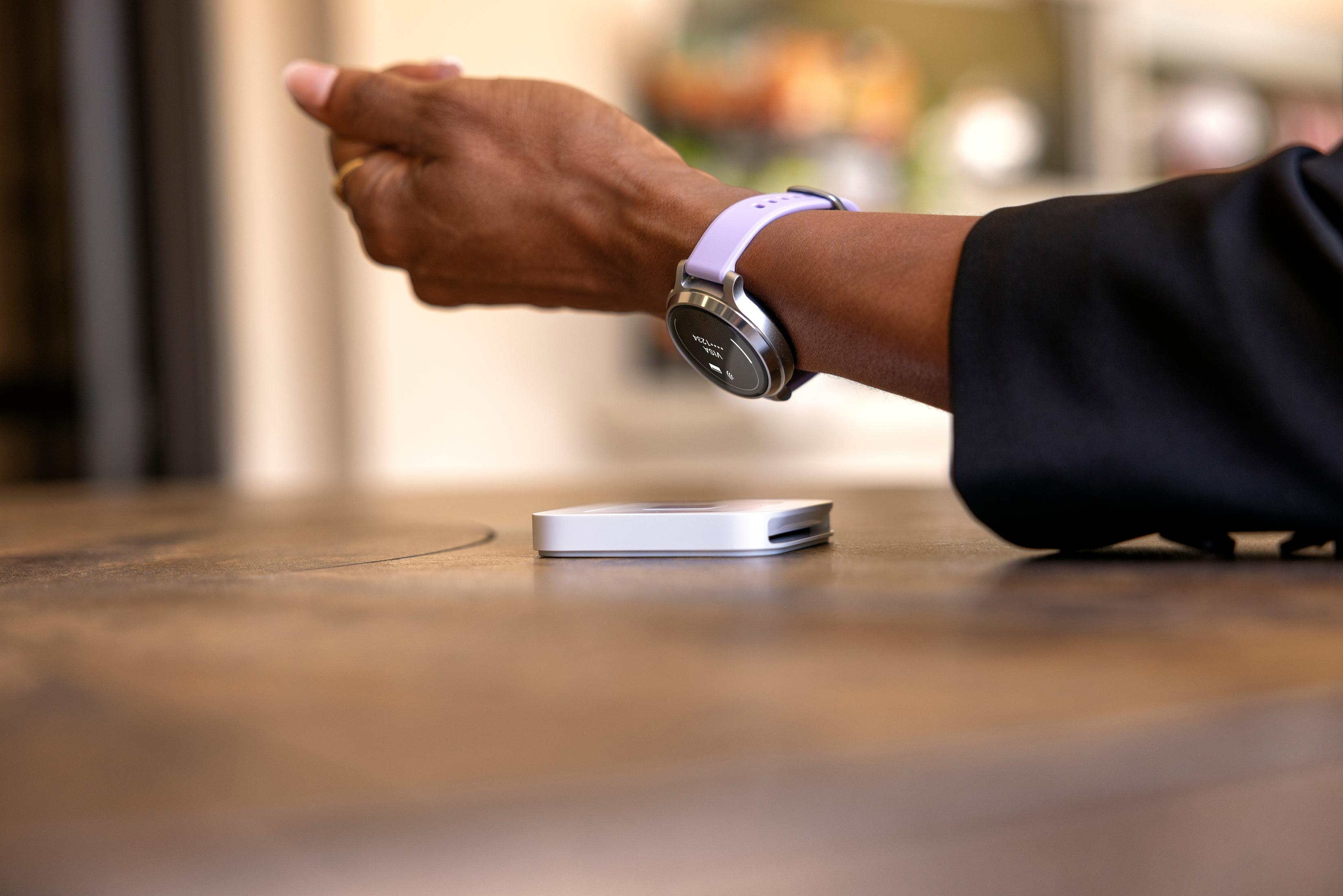 Eine Hand mit einer lila Smartwatch und einem weißen Gerät auf einem Holztisch.