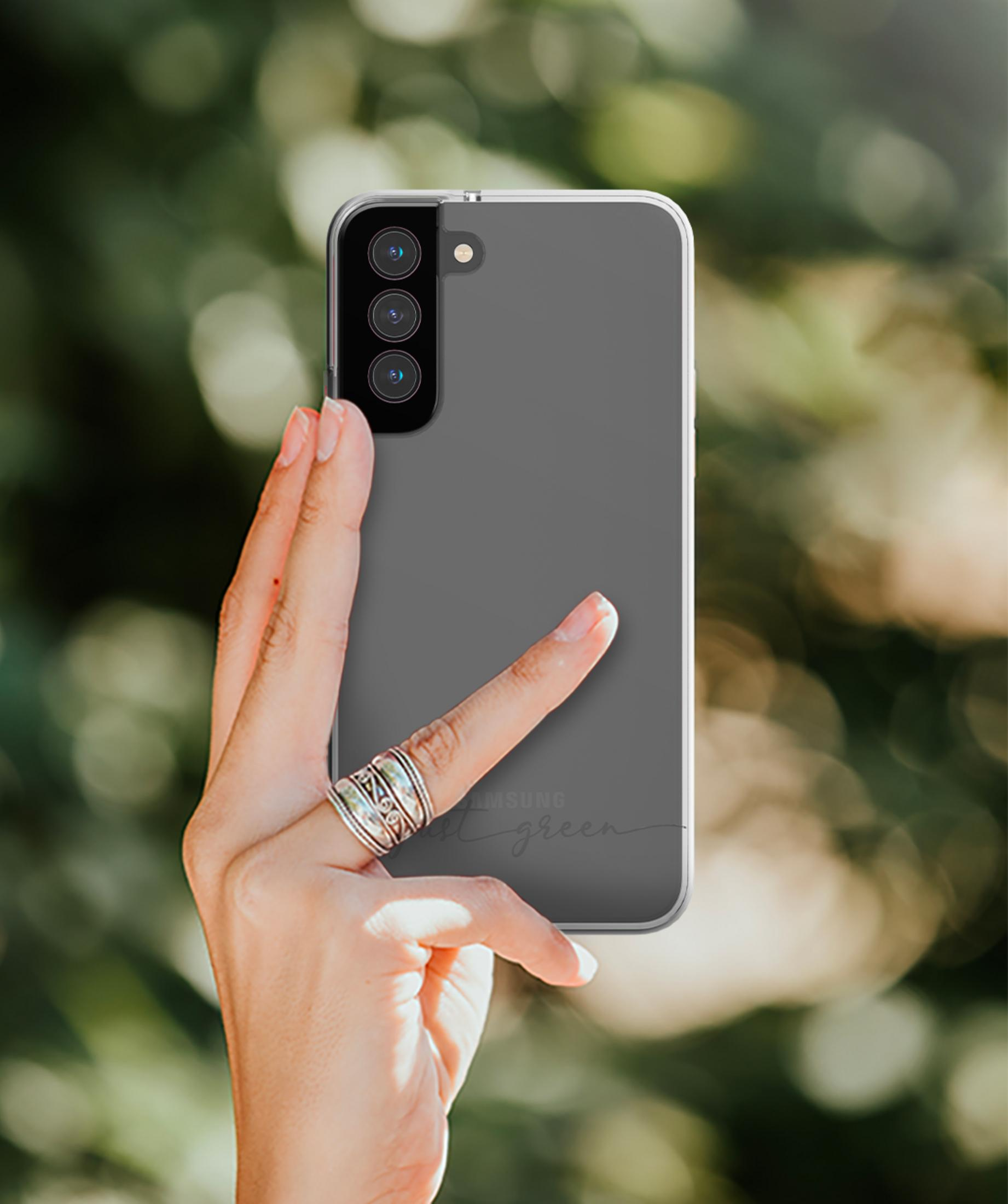 Hand hält ein transparentes Handy mit drei Kameras vor unscharfem grünen Hintergrund.