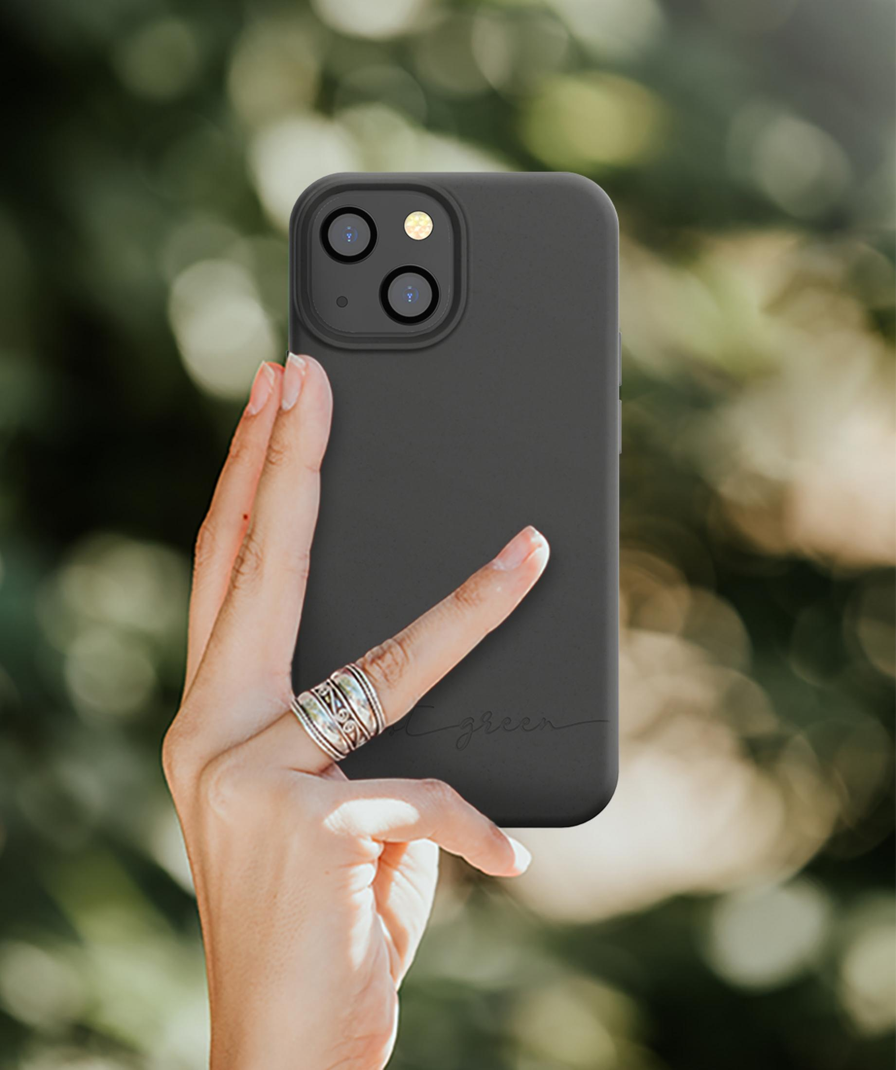 Una mano con anelli tiene una cover nera per telefono su sfondo verde sfocato.