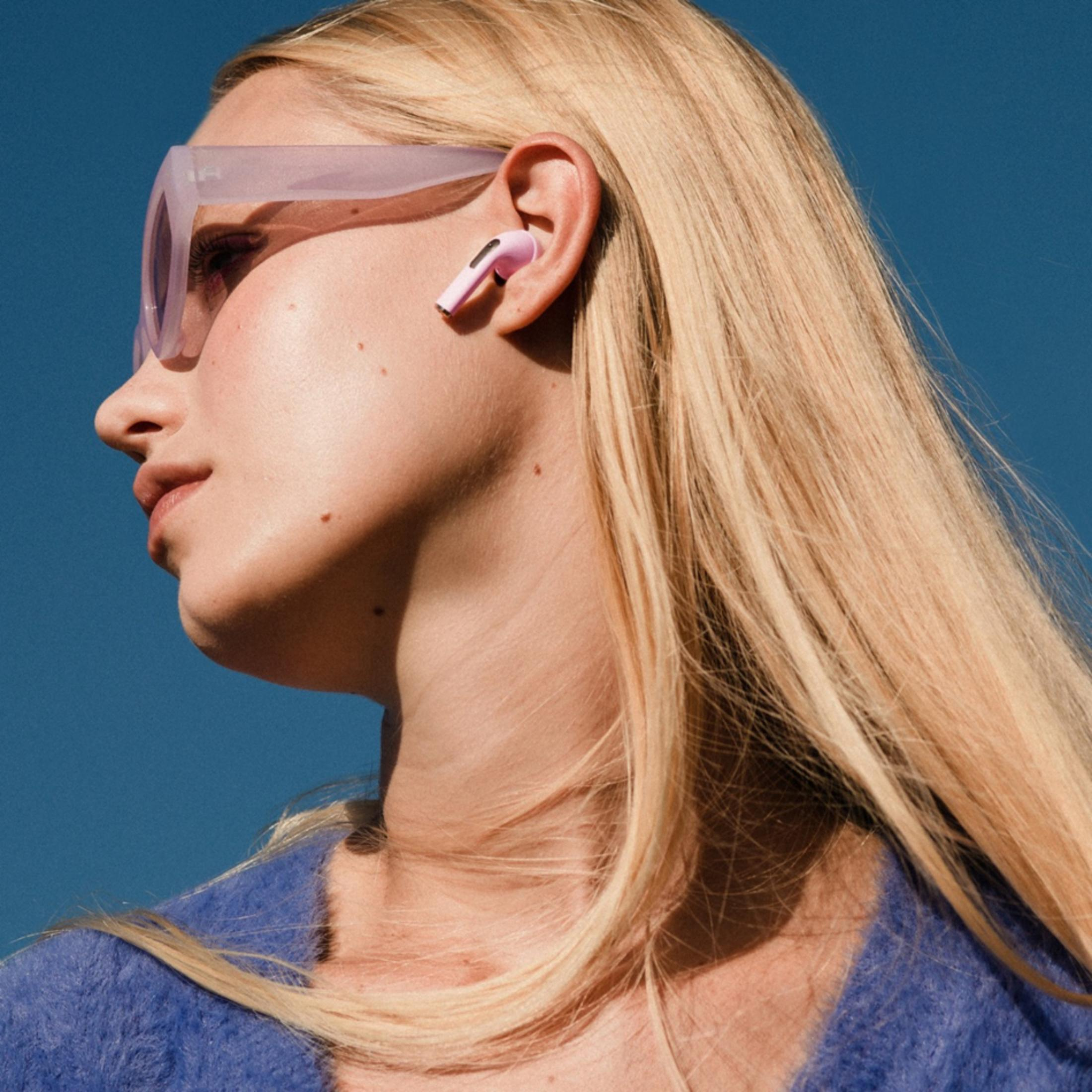 Una donna bionda con auricolari rosa e occhiali da sole su sfondo blu.