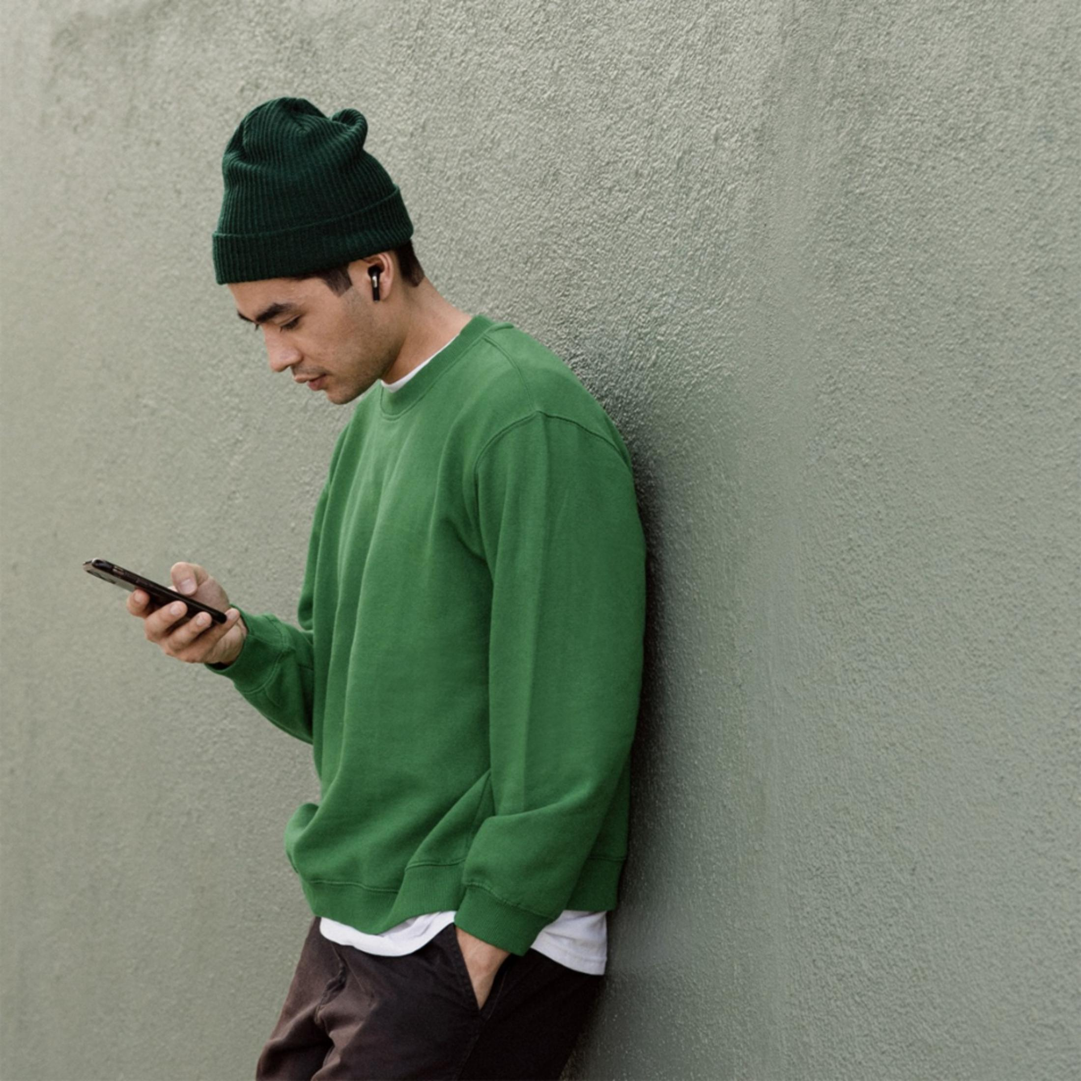 Ein Mann in grünen Kleidern lehnt an einer Wand und schaut auf sein Handy.