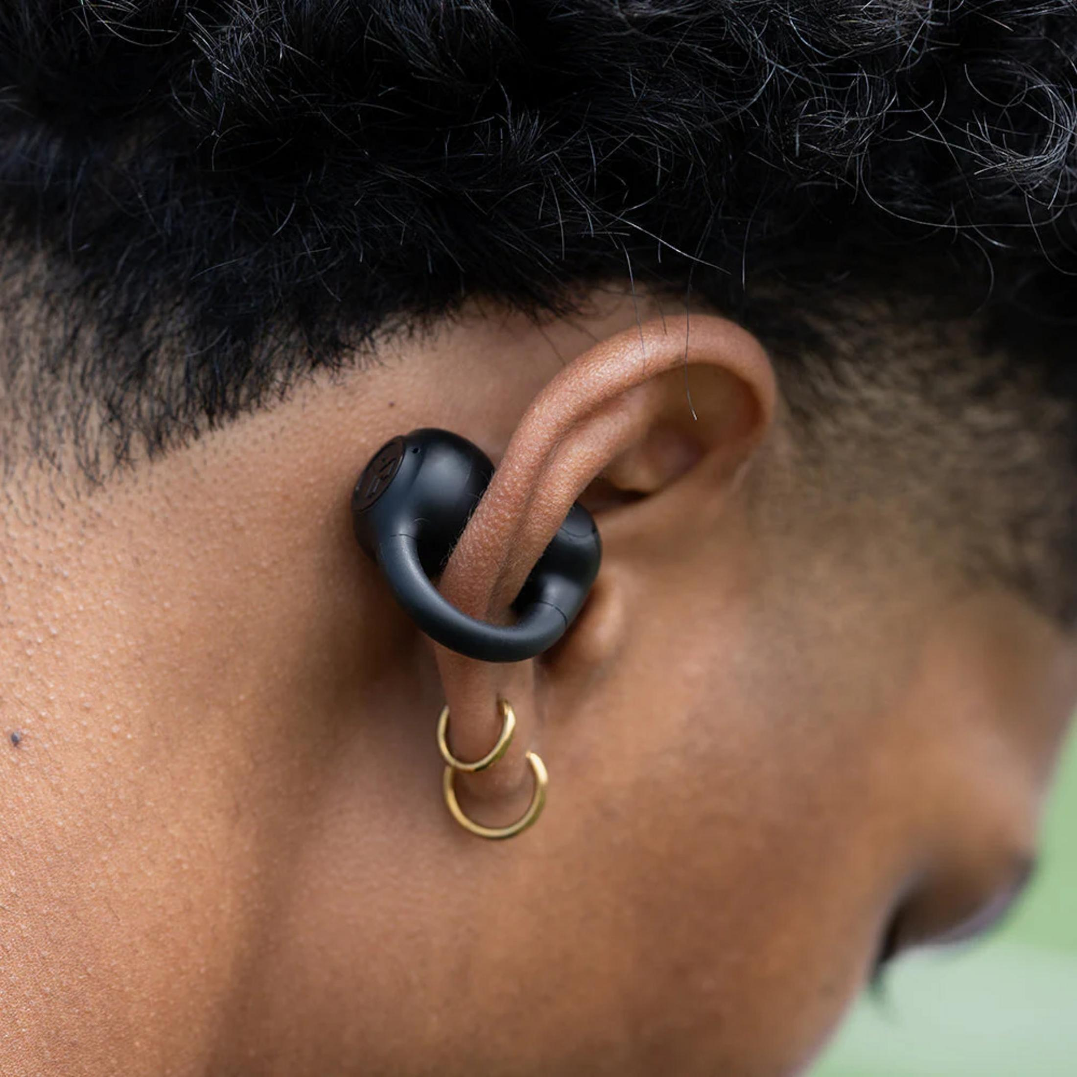 Un écouteur noir à crochet est porté dans l'oreille d'un homme. Des boucles d'oreilles dorées sont visibles.