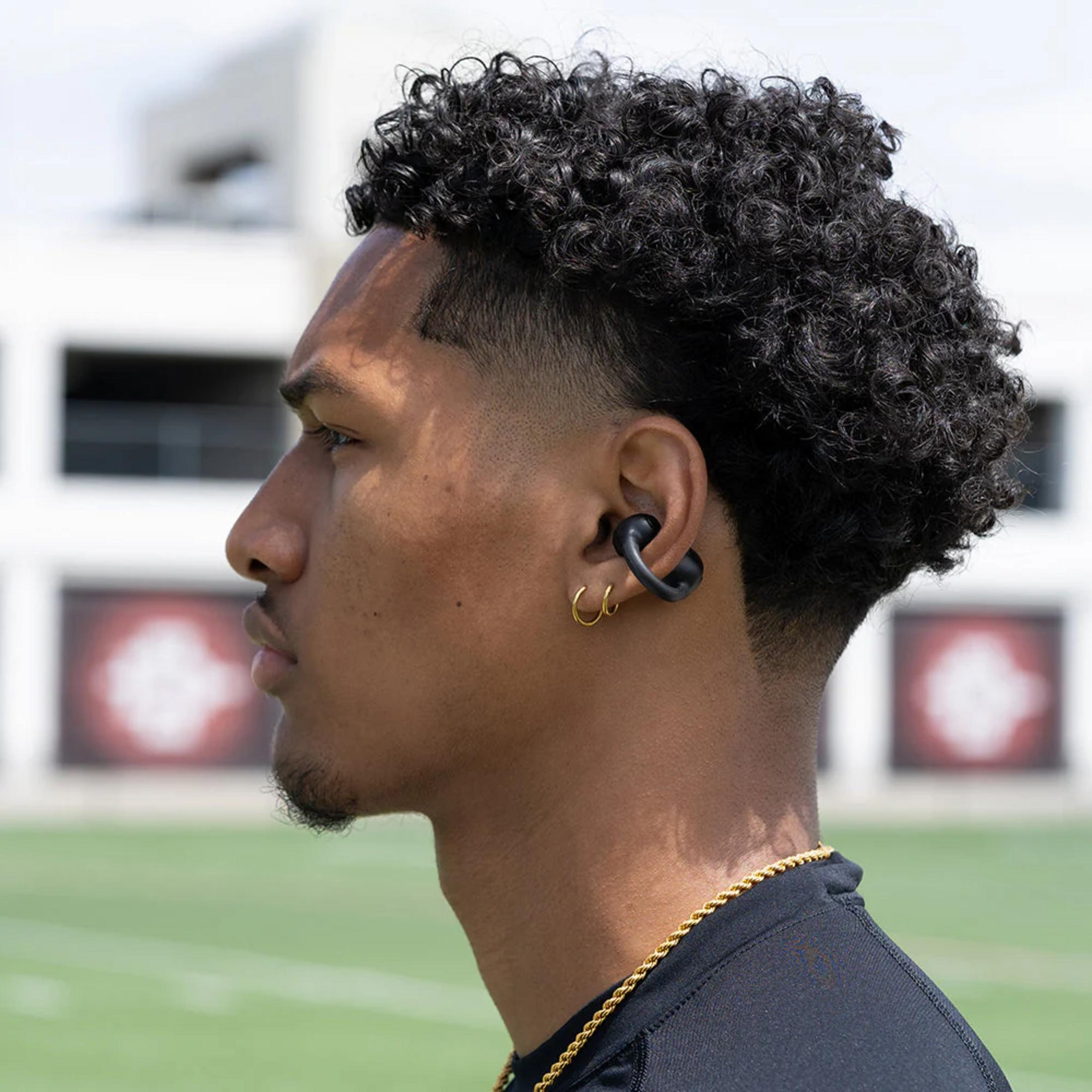 Homme aux cheveux bouclés, écouteur et chaîne en or sur un terrain de sport.