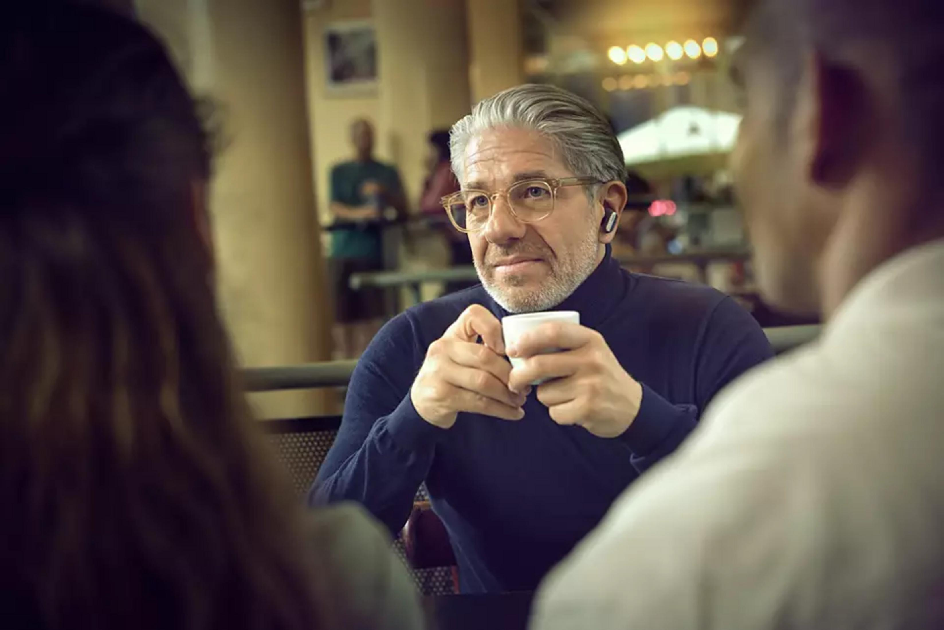 Un uomo con gli occhiali beve un caffè. Indossa un maglione a collo alto blu.