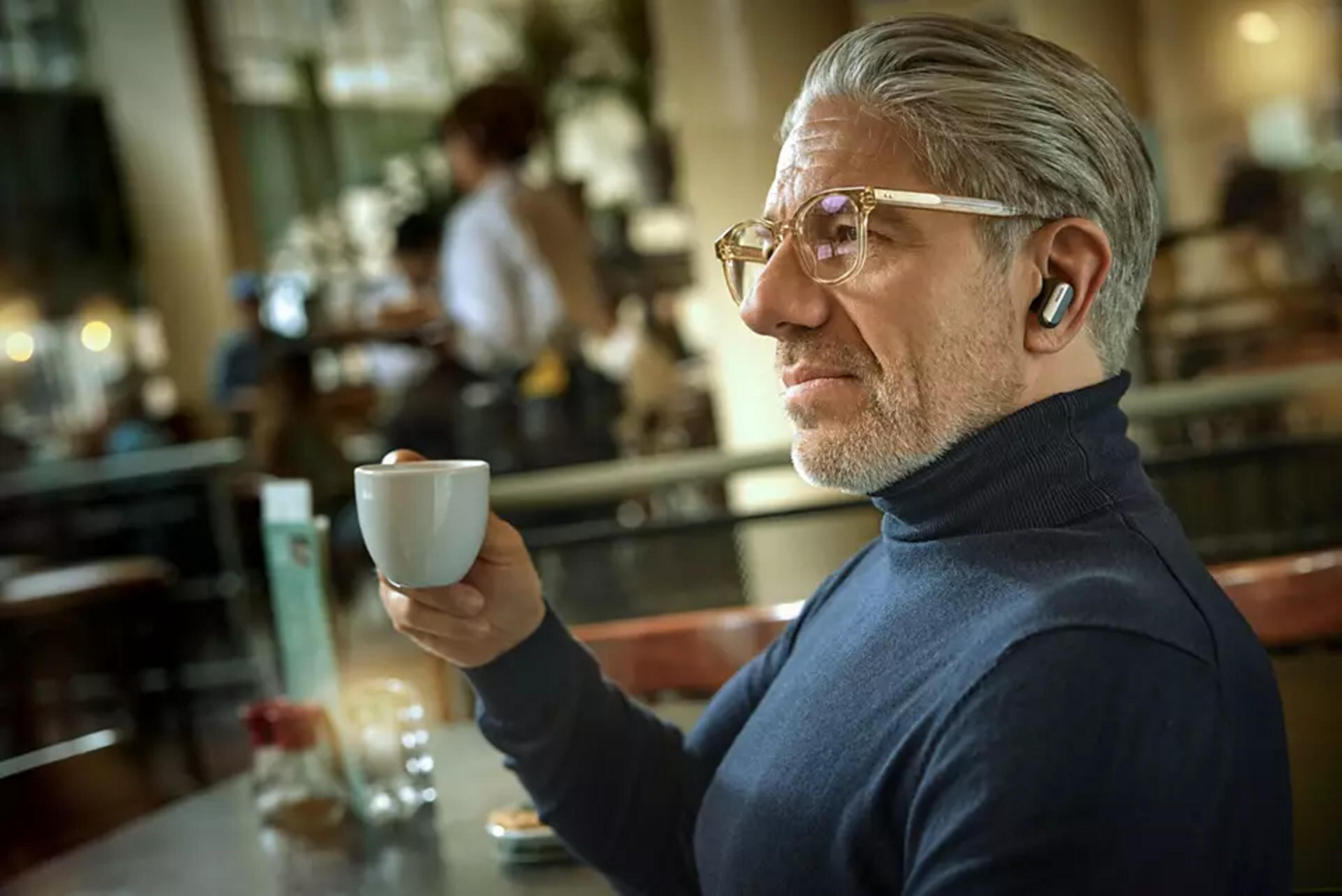 Un uomo con occhiali e auricolare tiene una tazza di caffè in un bar.
