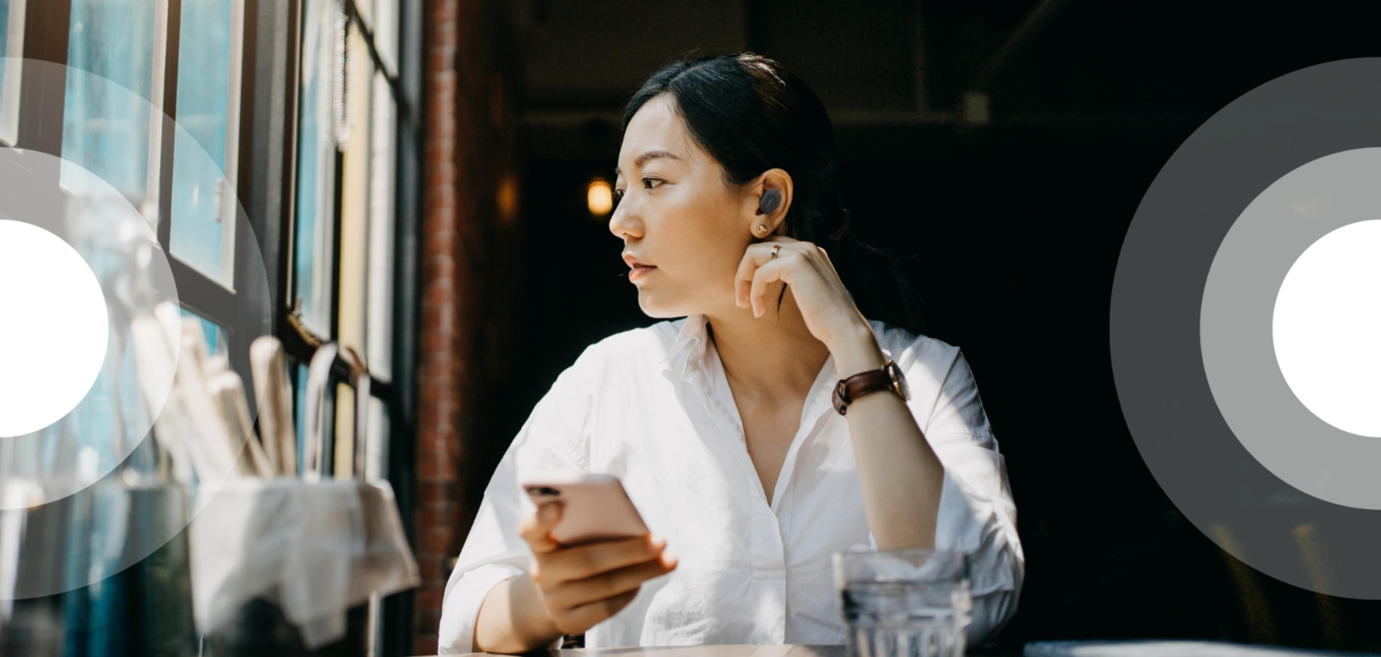 Kobieta ze słuchawkami trzyma telefon i patrzy przez okno.