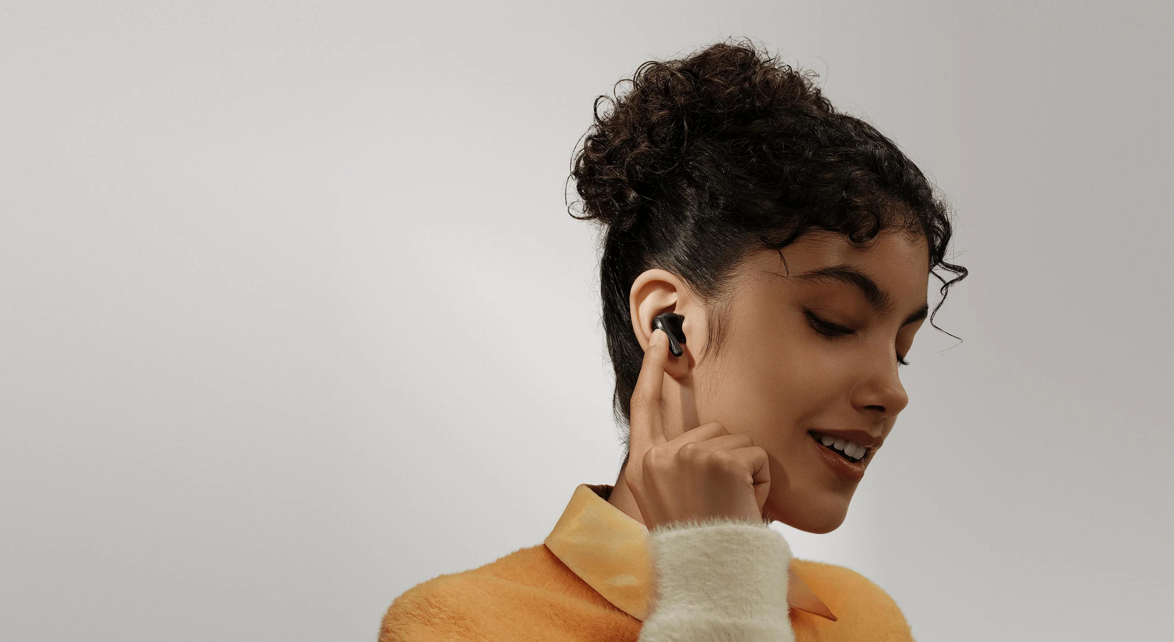 Kobieta z ciemnymi kręconymi włosami nosi pomarańczowy sweter i czarny słuchawkę.