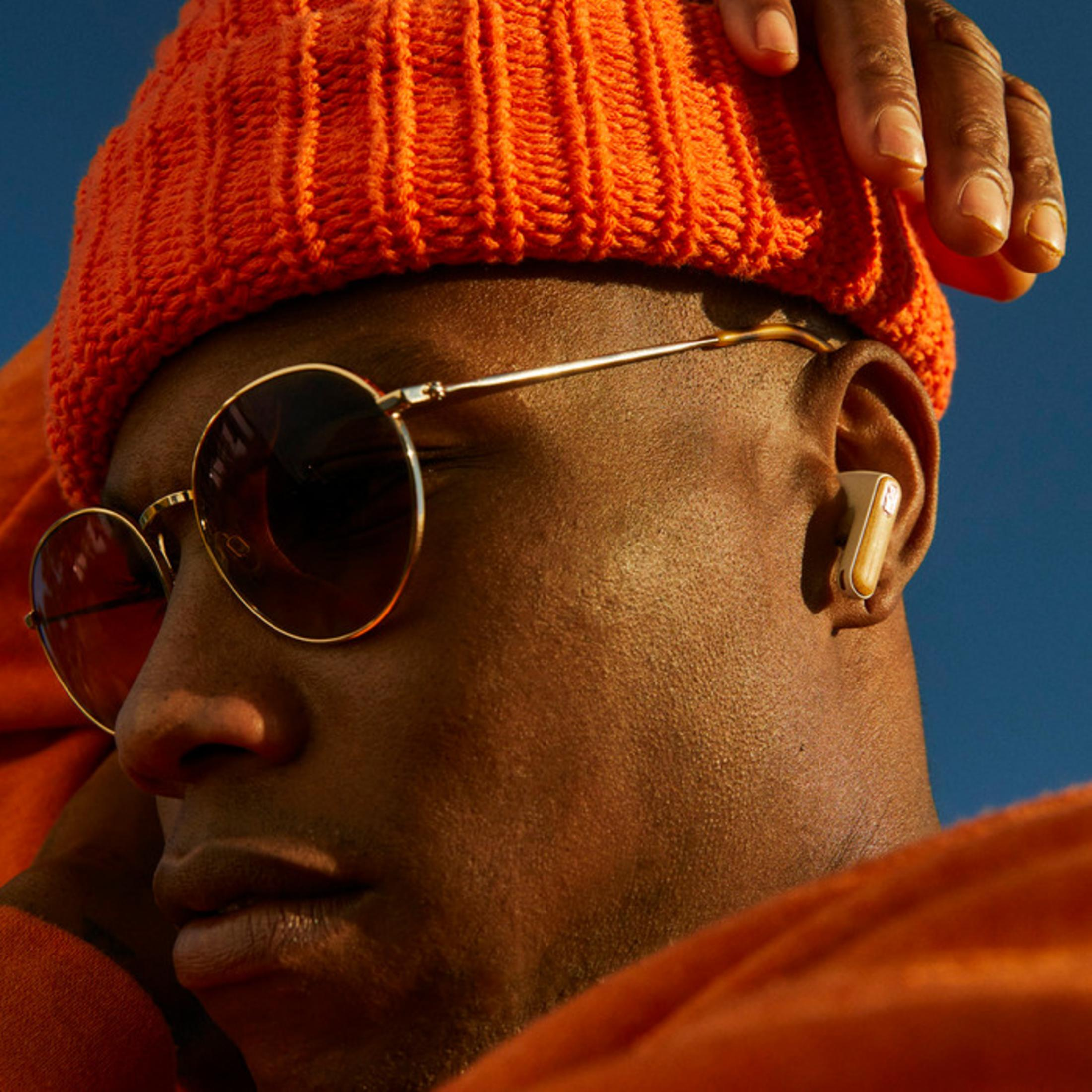 Mann mit orangener Mütze, Sonnenbrille und Ohrhörer vor blauem Himmel.