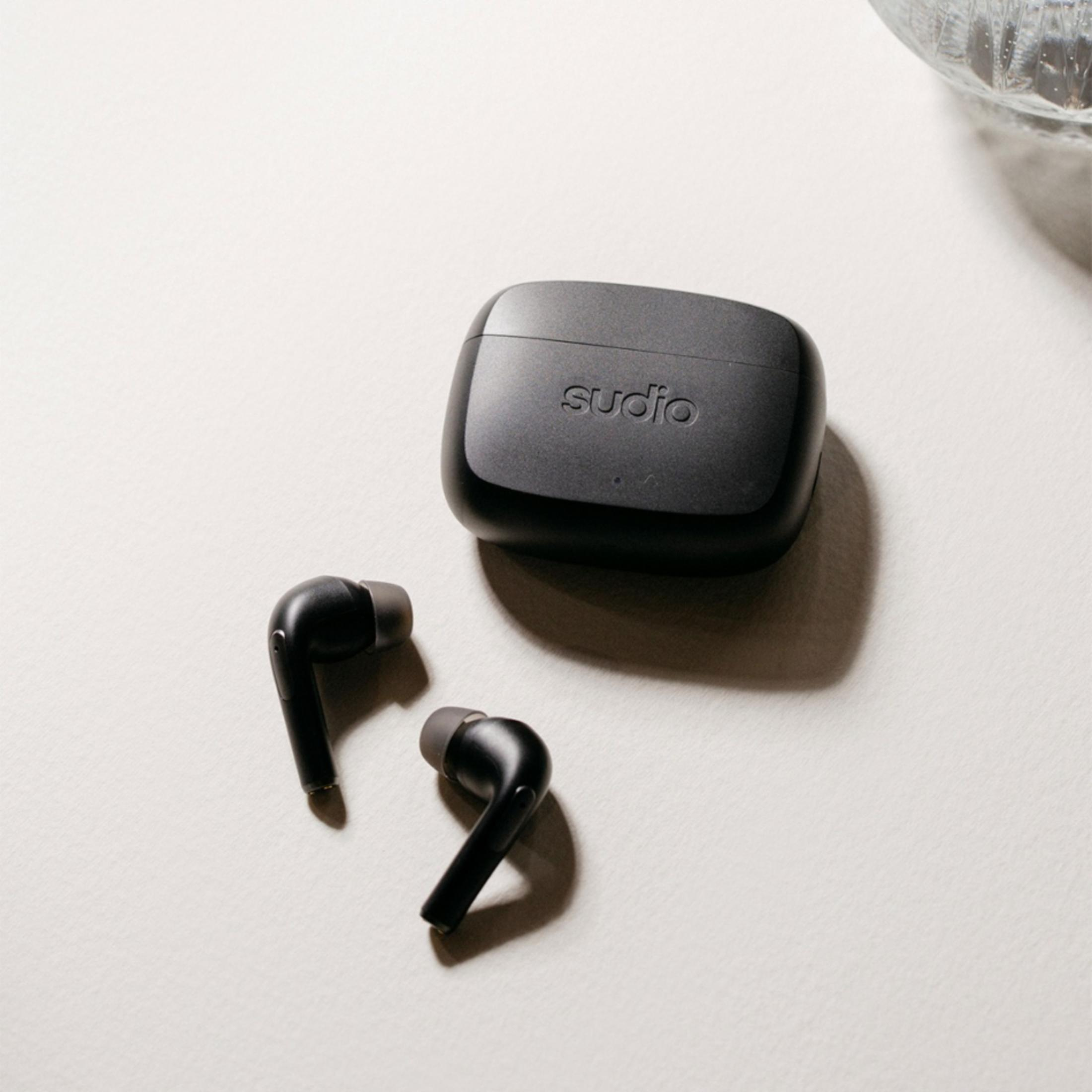 Custodia di ricarica Sudio nera con due auricolari wireless accanto su superficie chiara.