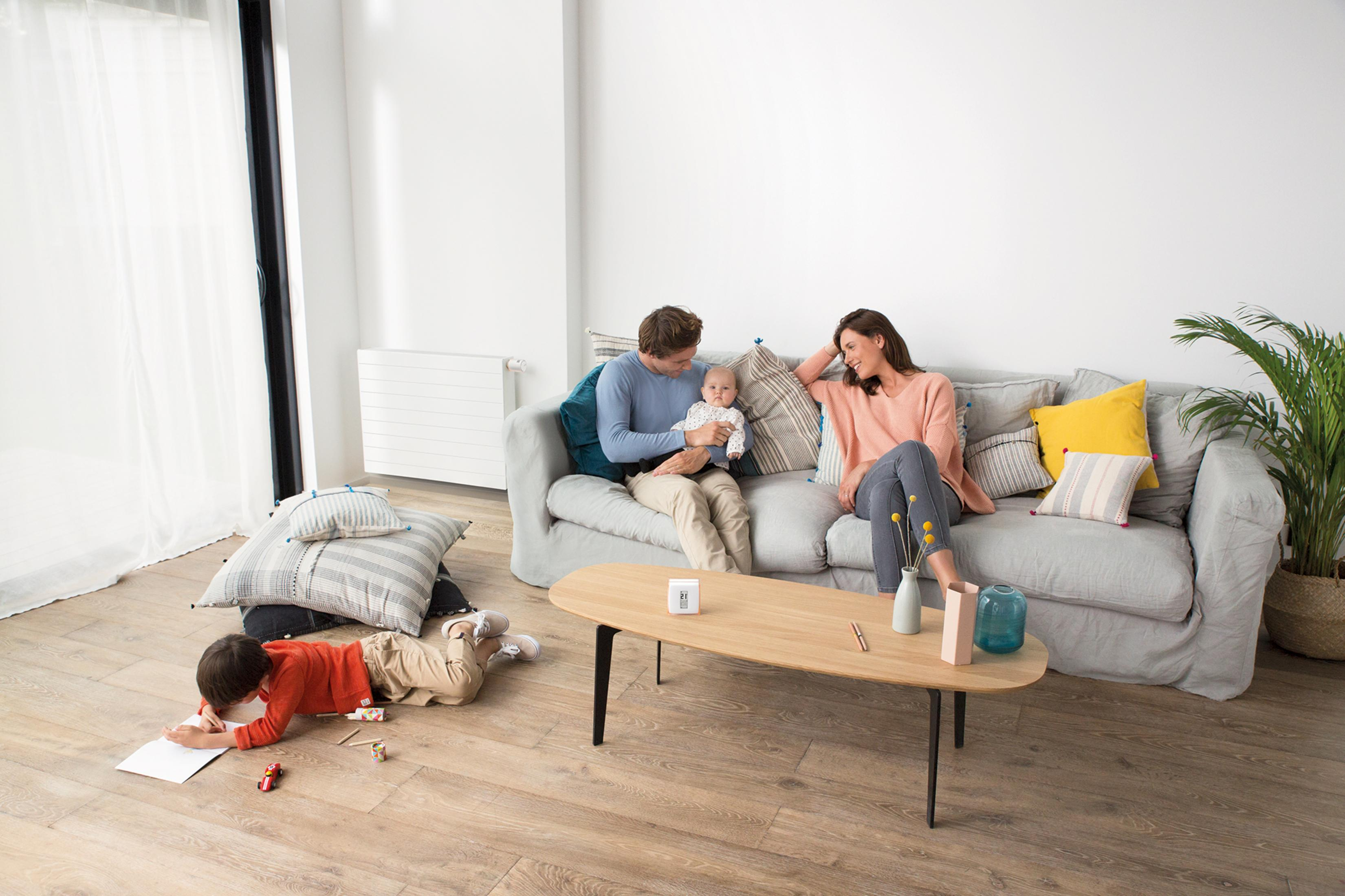 Familie im Wohnzimmer: Vater hält Baby, Mutter schaut zu, Kind malt am Boden.