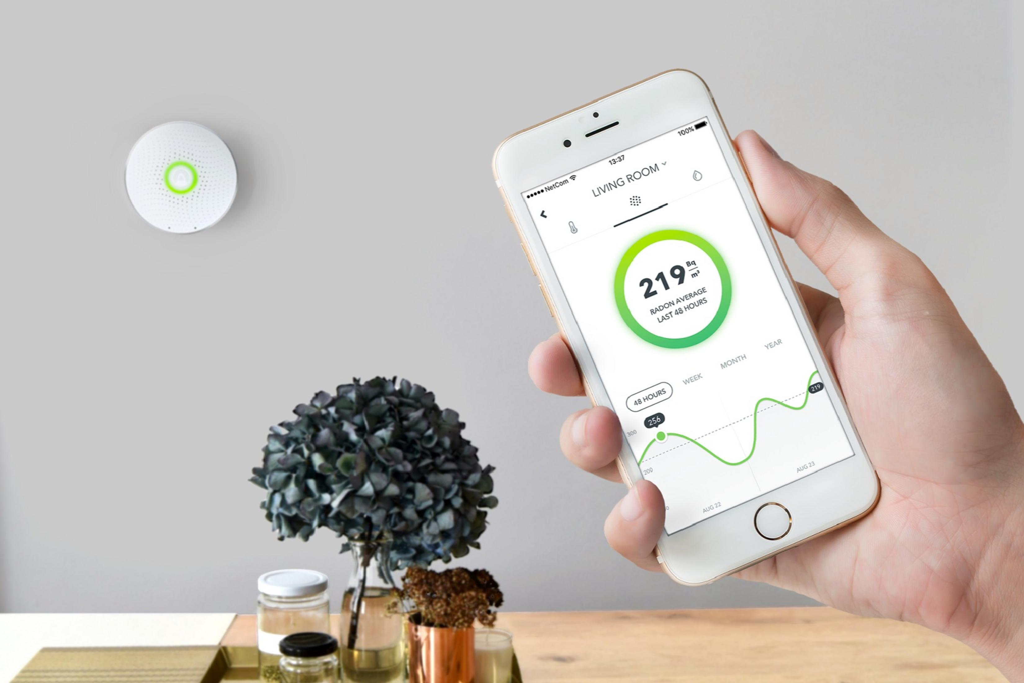 Ein weißer Radonmelder hängt an der Wand. Eine Hand hält ein Smartphone mit Radonwerten.