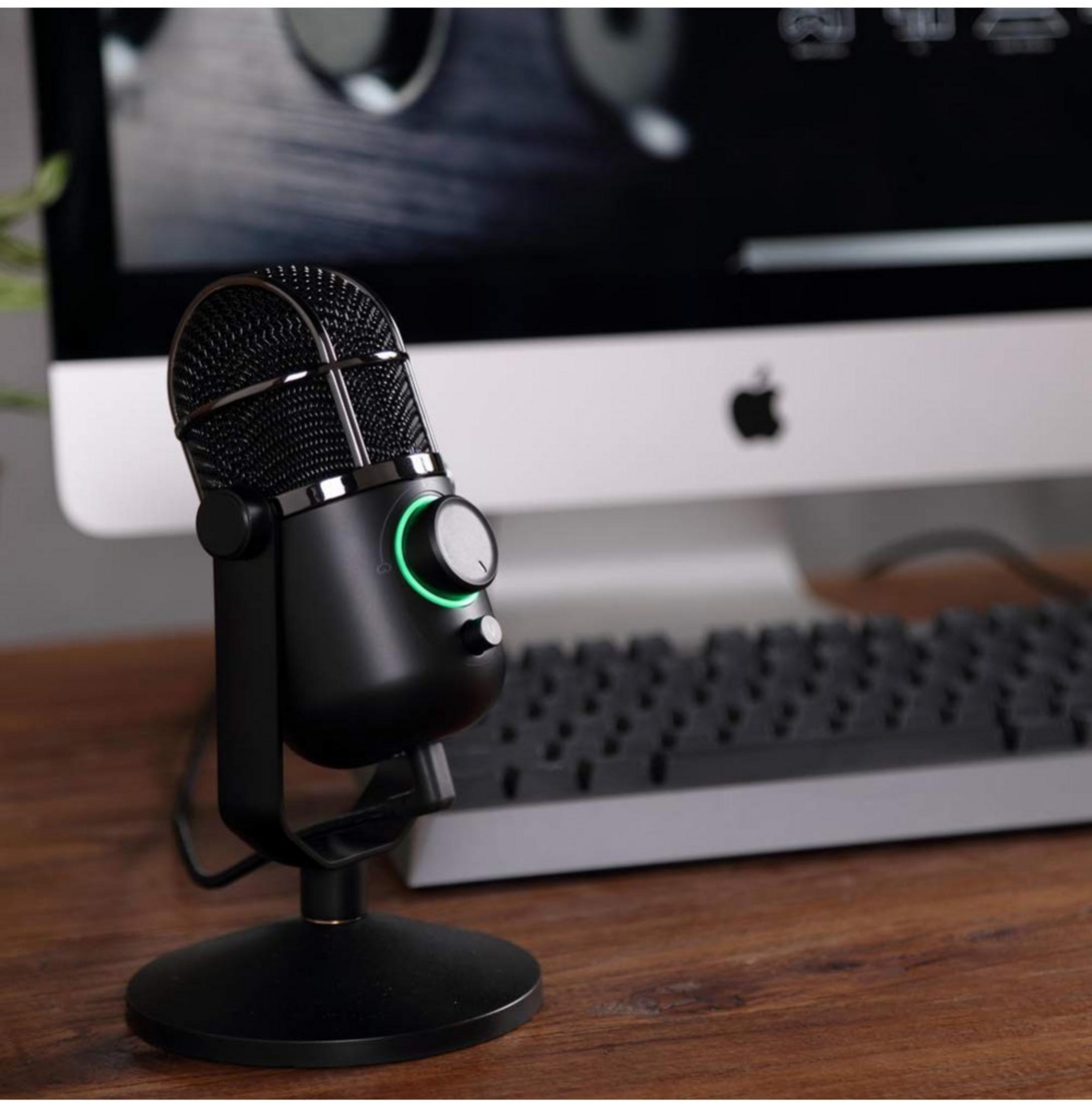 Un microfono nero con luce verde è su una scrivania di legno, accanto a una tastiera e un computer Mac.