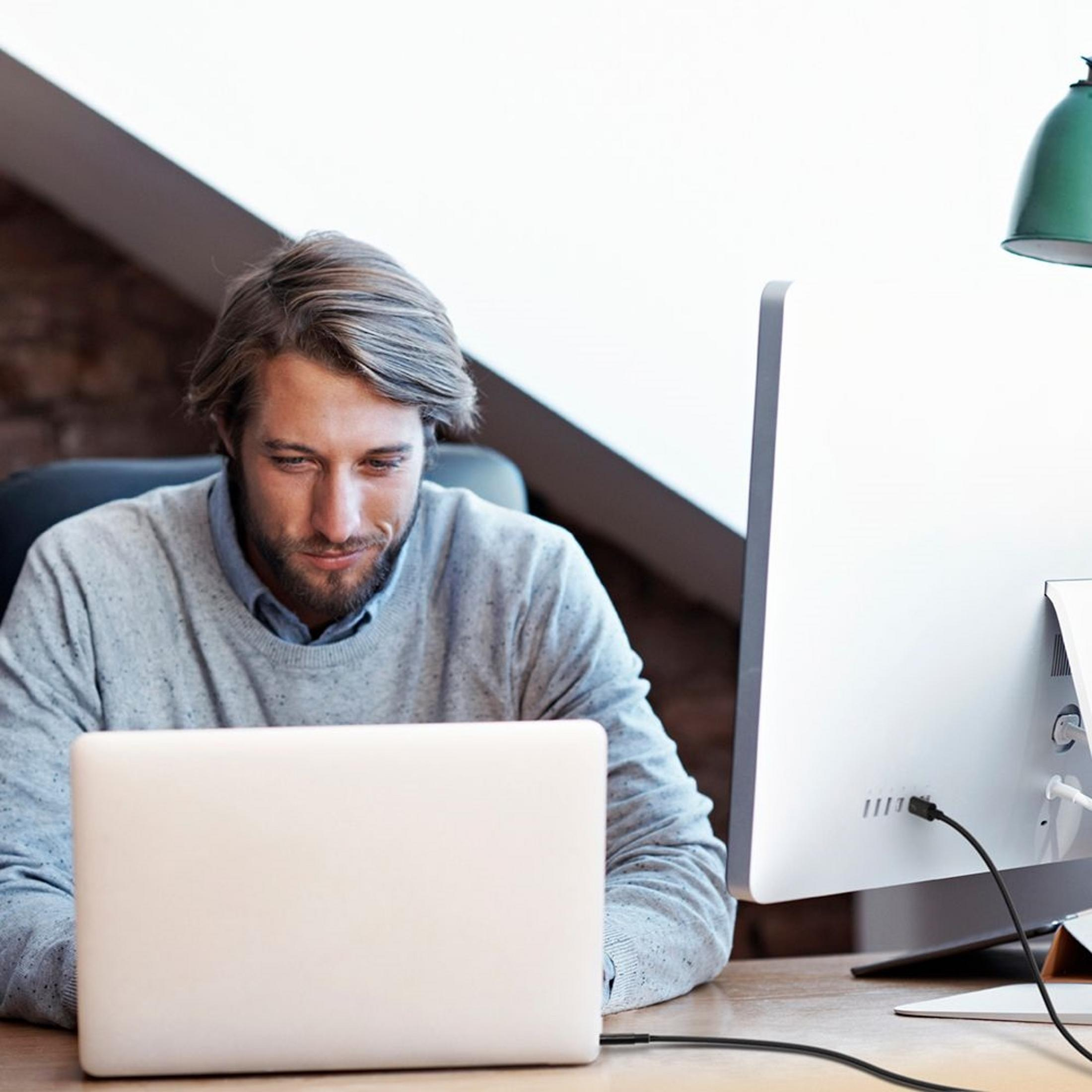 Ein Mann sitzt an einem Schreibtisch und arbeitet an einem Laptop und einem Computer.