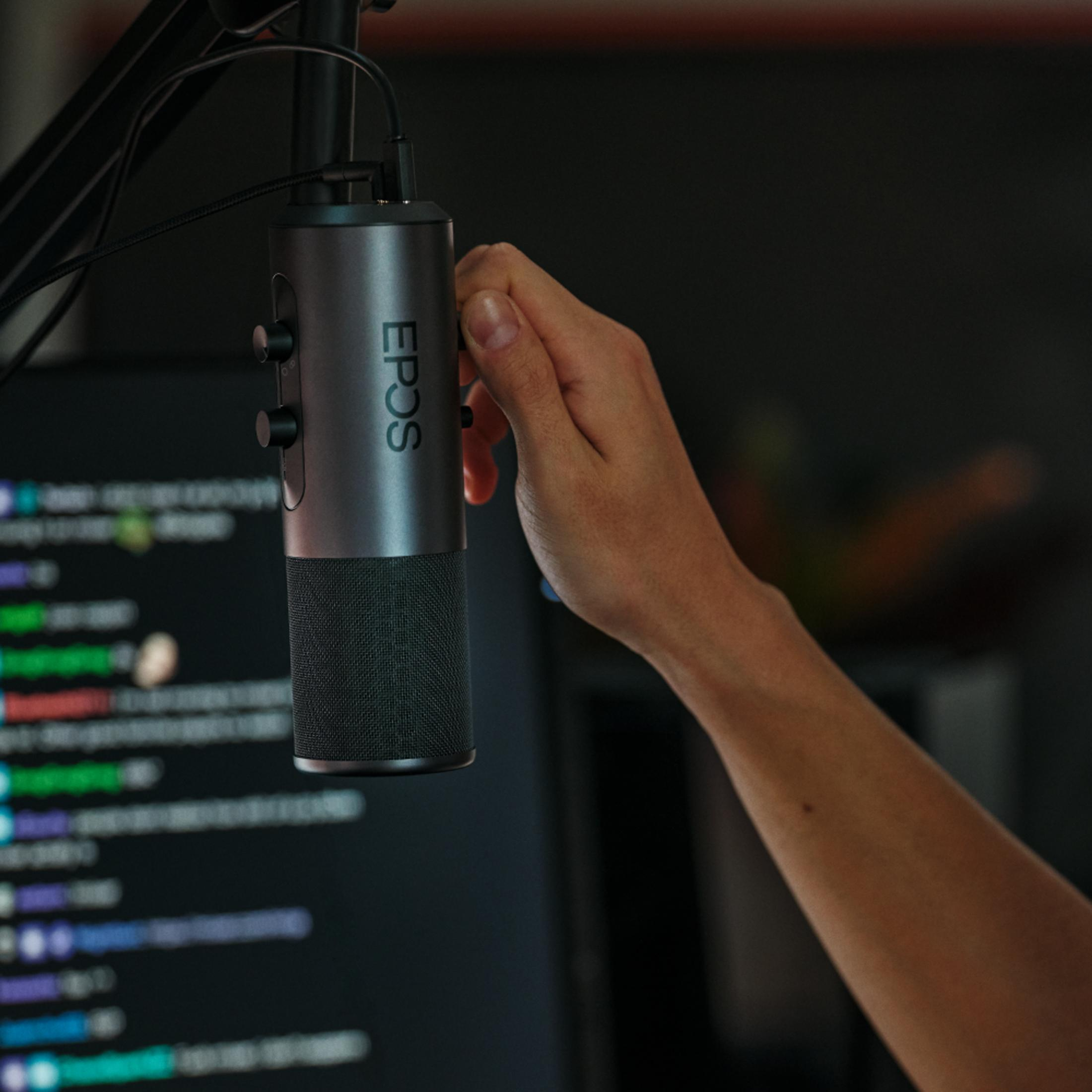 Eine Hand dreht an einem EPOS-Mikrofon vor einem Computerbildschirm mit Code.
