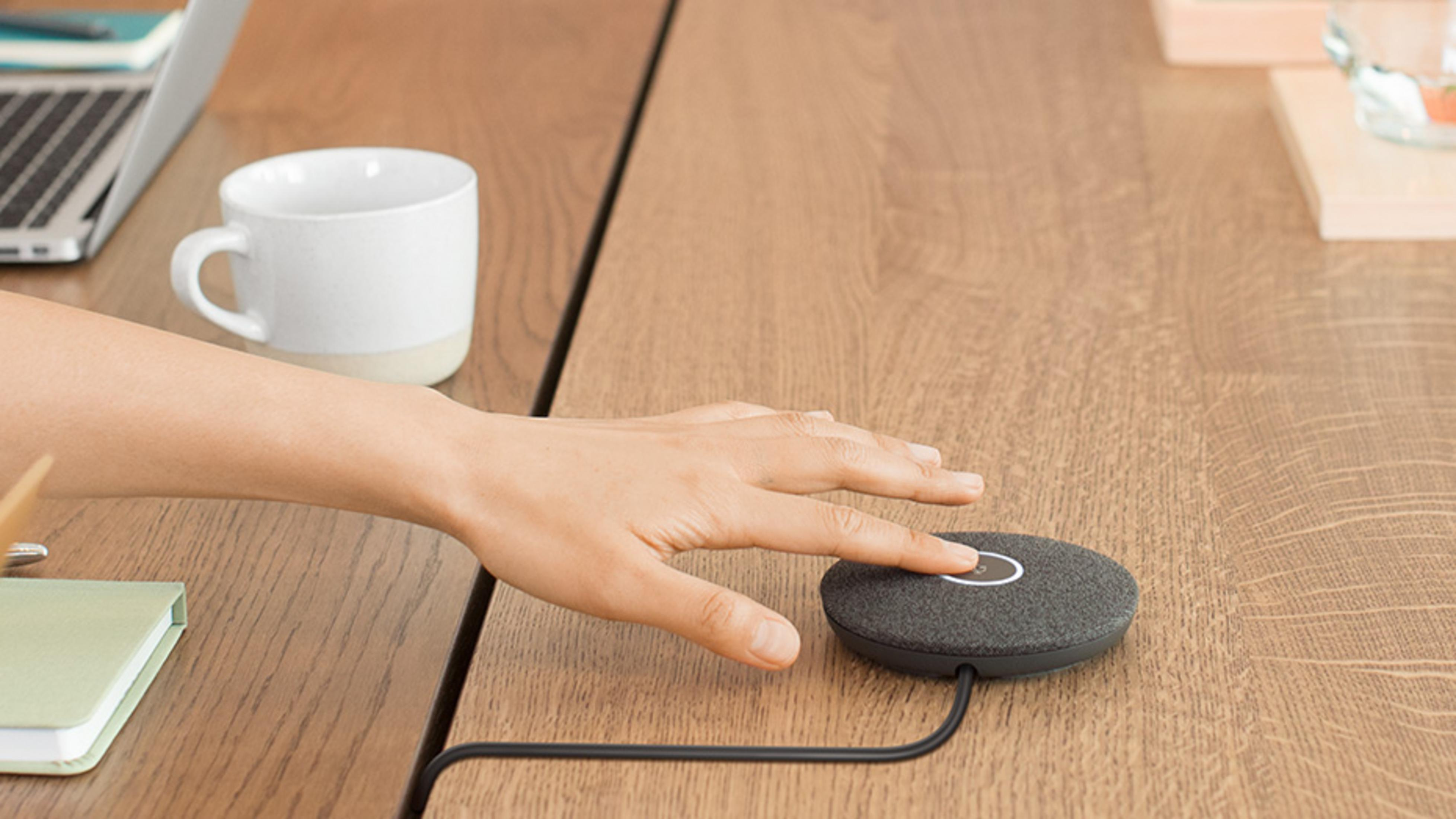 Dłoń dotyka szarego mikrofonu na drewnianym stole obok kubka i laptopa.