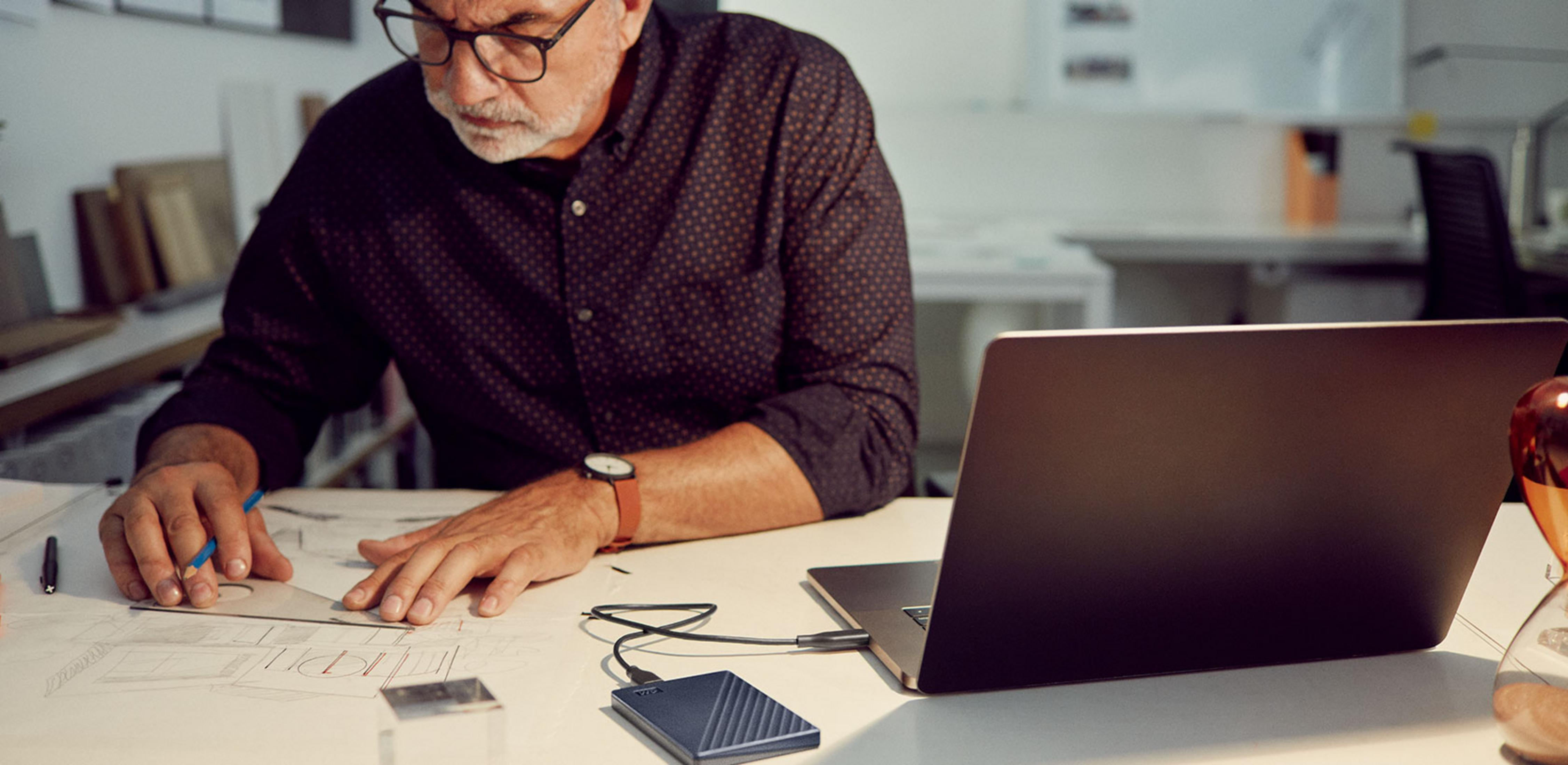 Mężczyzna w okularach rysuje plany z dyskiem zewnętrznym i laptopem.