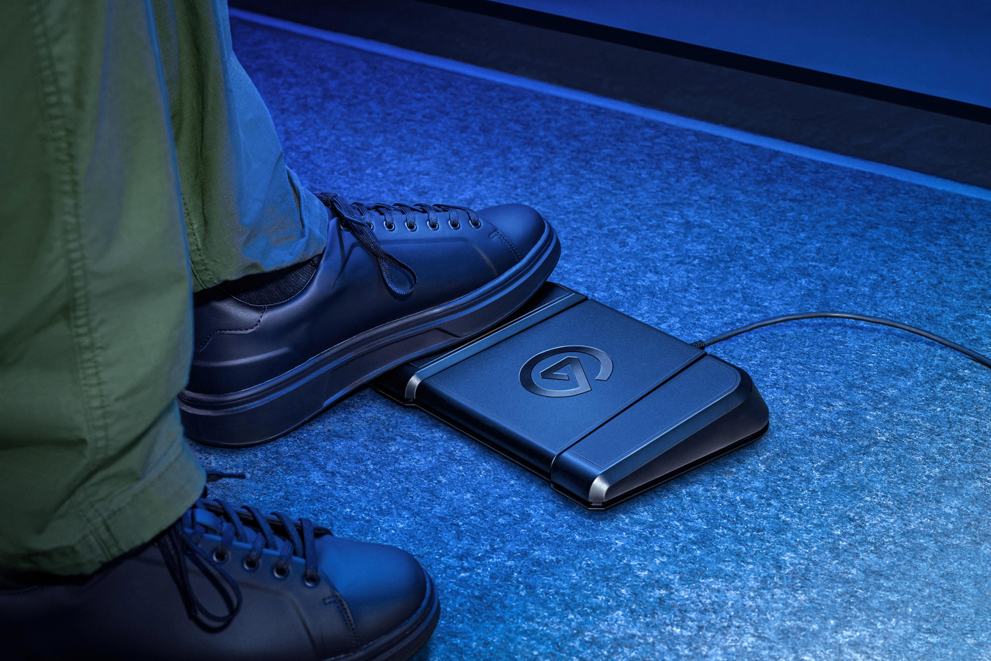 Fuß auf schwarzem Pedal mit Logo, grüne Hose, blaue Beleuchtung.