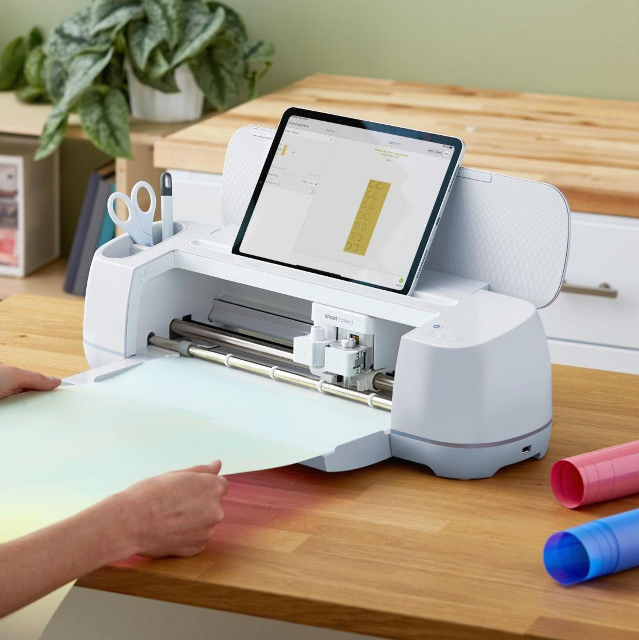 Eine weiße Cricut Maker 3 Maschine mit Tablet steht auf einem Holztisch. Eine Hand legt ein Materialblatt in die Maschine.