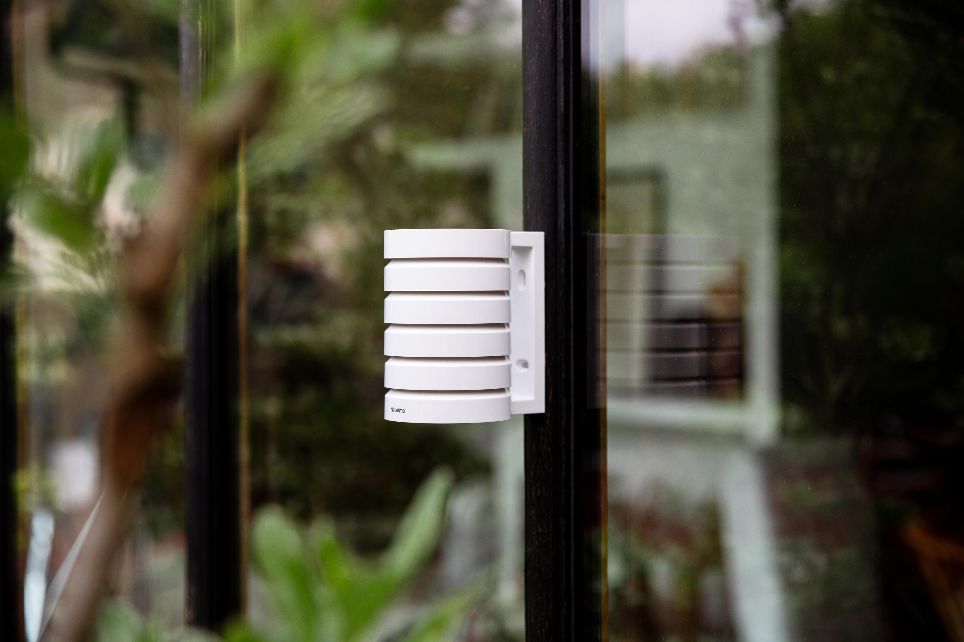 A white Netatmo device with horizontal slats is attached to a dark window frame.