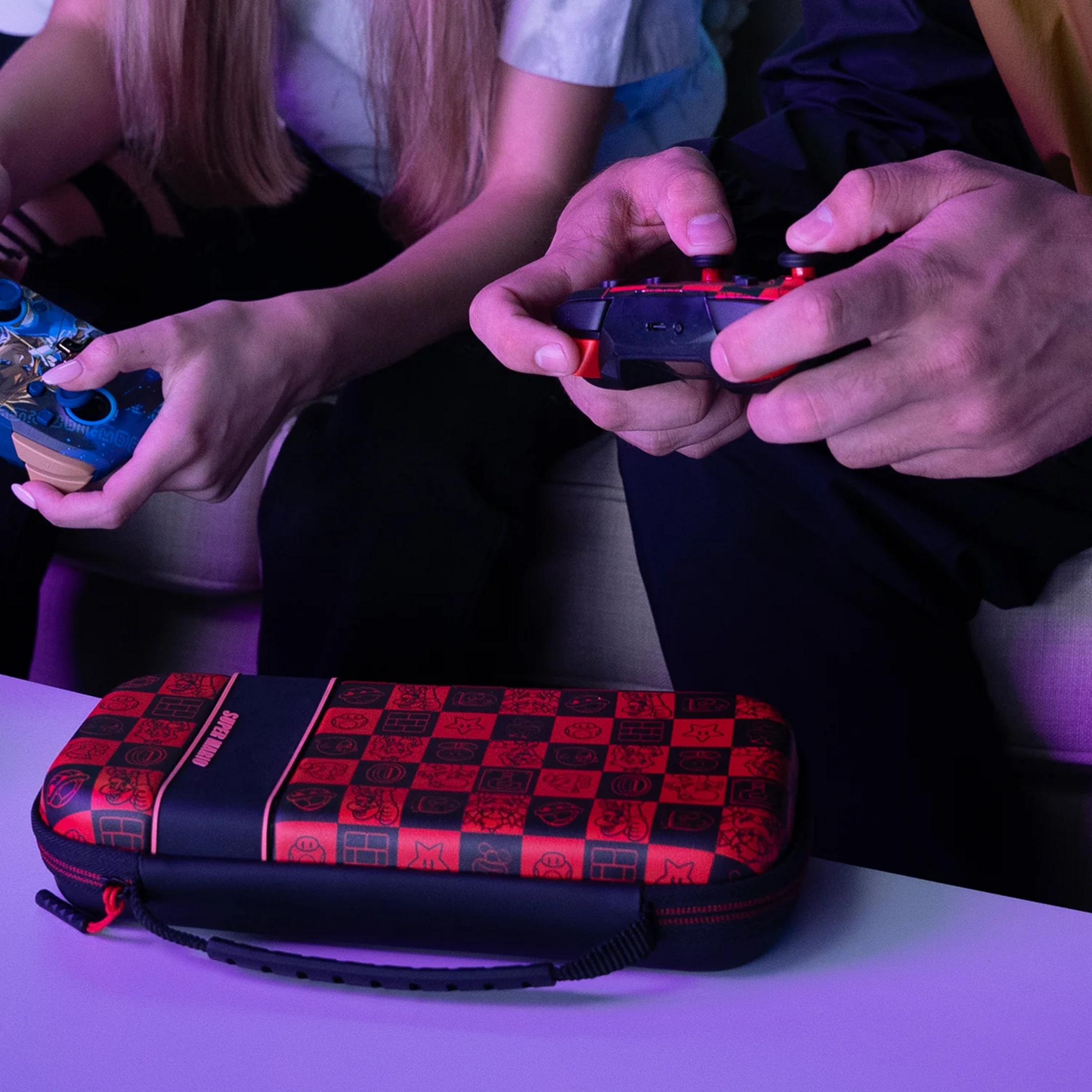 Dwie osoby grają w gry wideo z kontrolerami. Z przodu znajduje się etui z motywem Mario.
