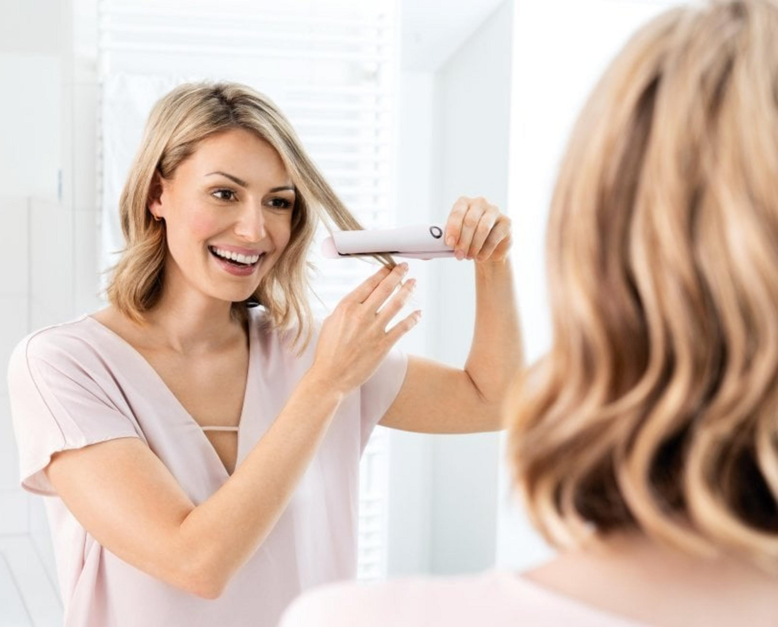 Eine lächelnde Frau mit blonden Haaren glättet ihr Haar mit einem Glätteisen.