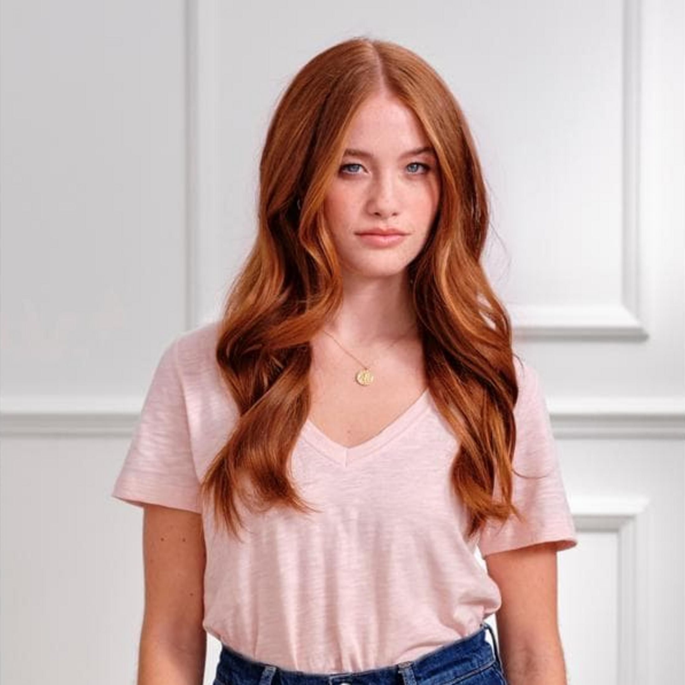 Eine junge Frau mit roten Haaren und blauen Augen trägt ein rosa T-Shirt.