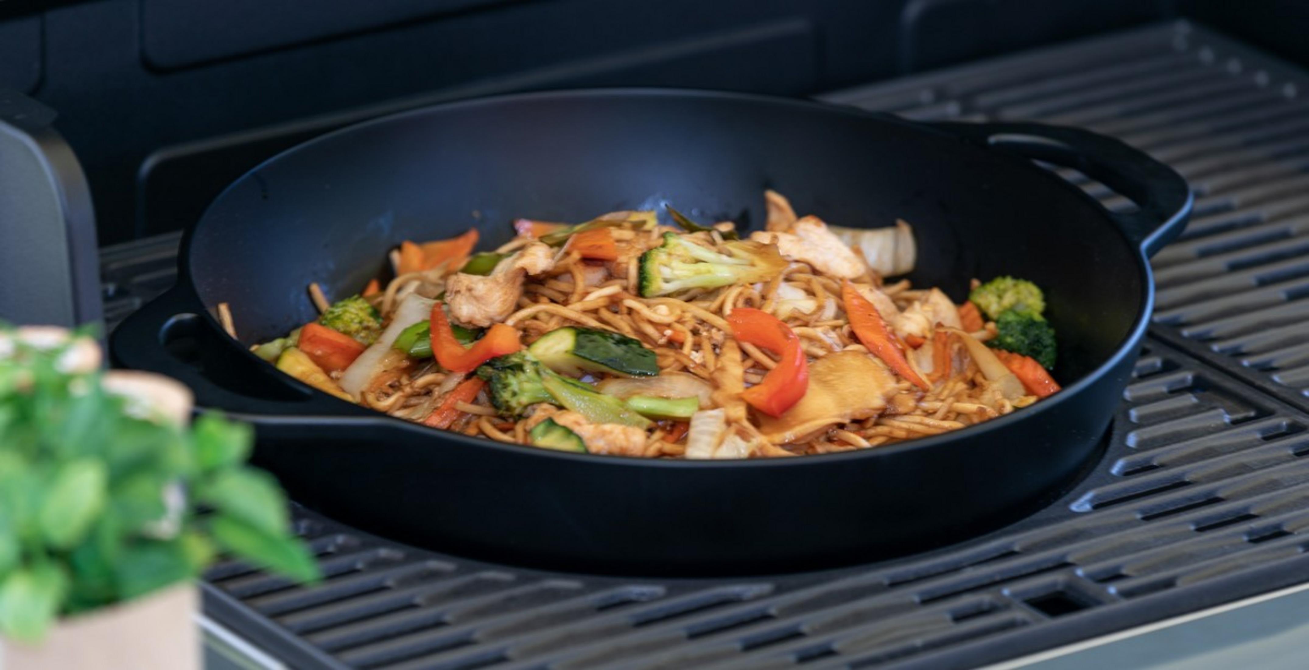 Un wok con fideos, pollo y verduras sobre una parrilla.