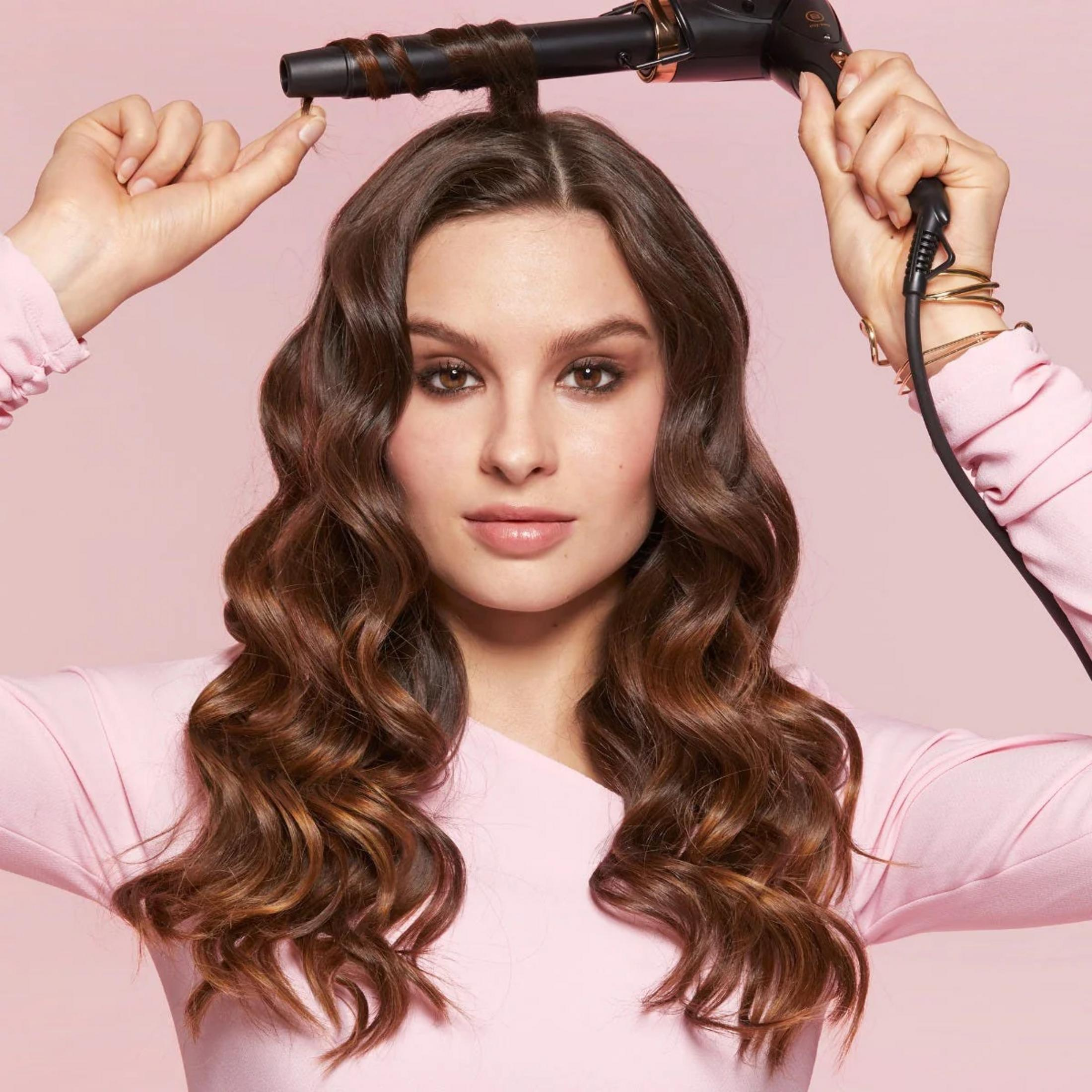 Mujer con rizador peinando su cabello sobre fondo rosa.
