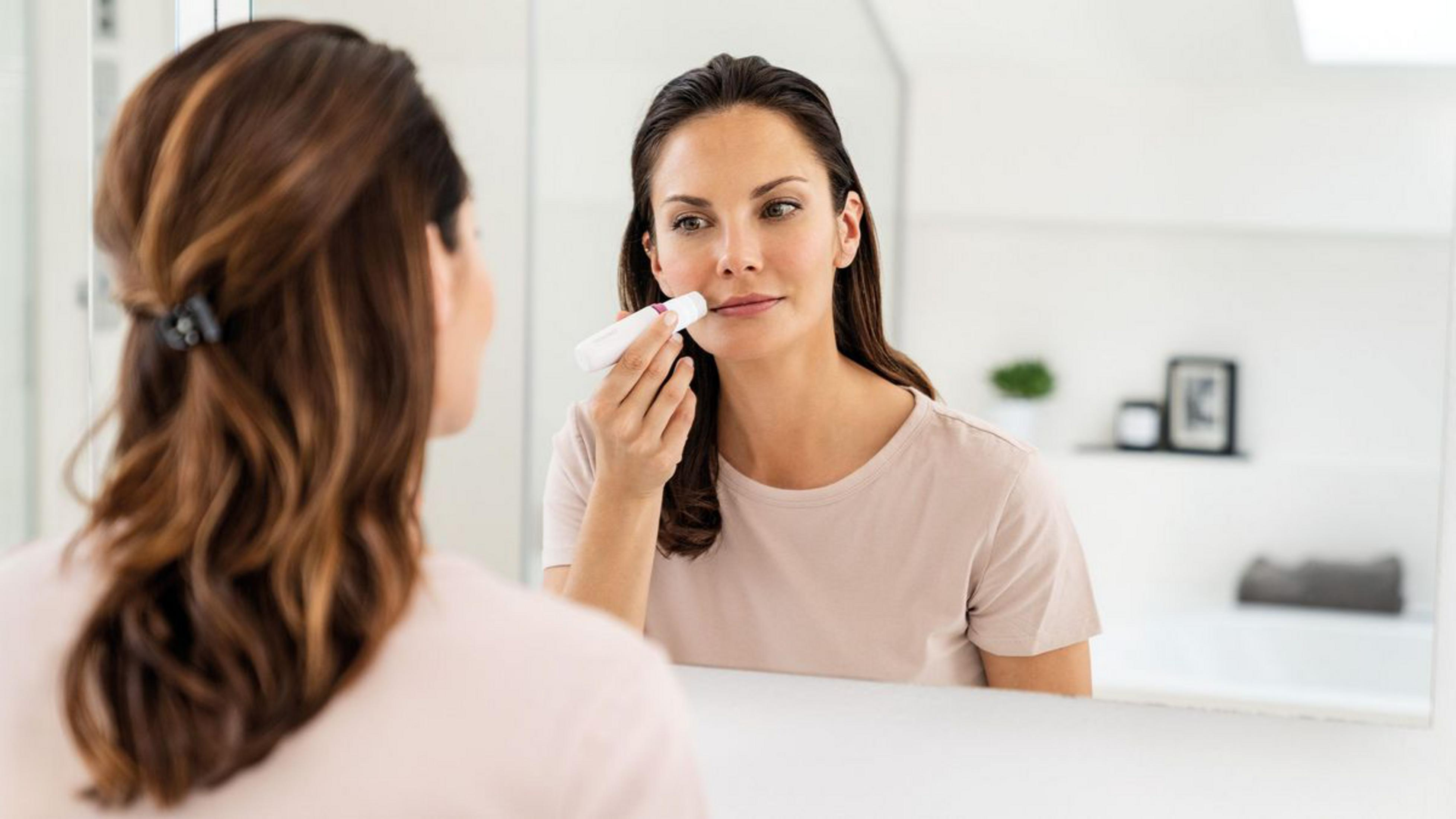 Mujer usa dispositivo de limpieza facial en baño, reflejada en espejo.