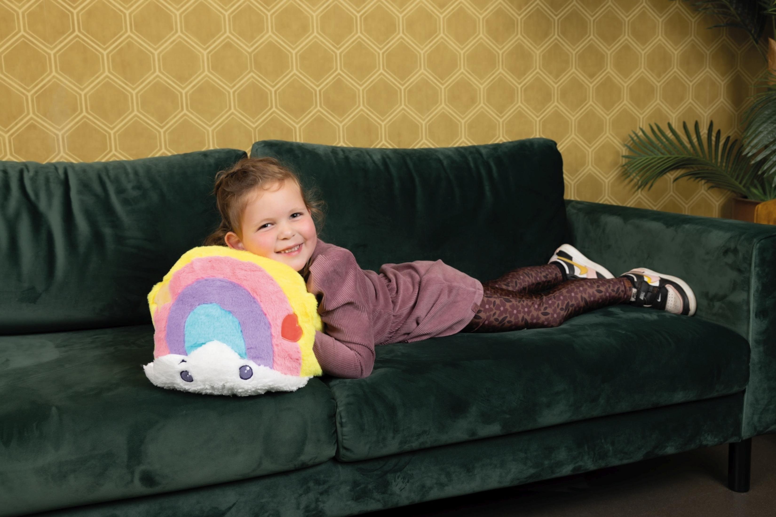 Ein lächelndes Mädchen liegt auf einer grünen Couch neben einem bunten Regenbogenkissen.