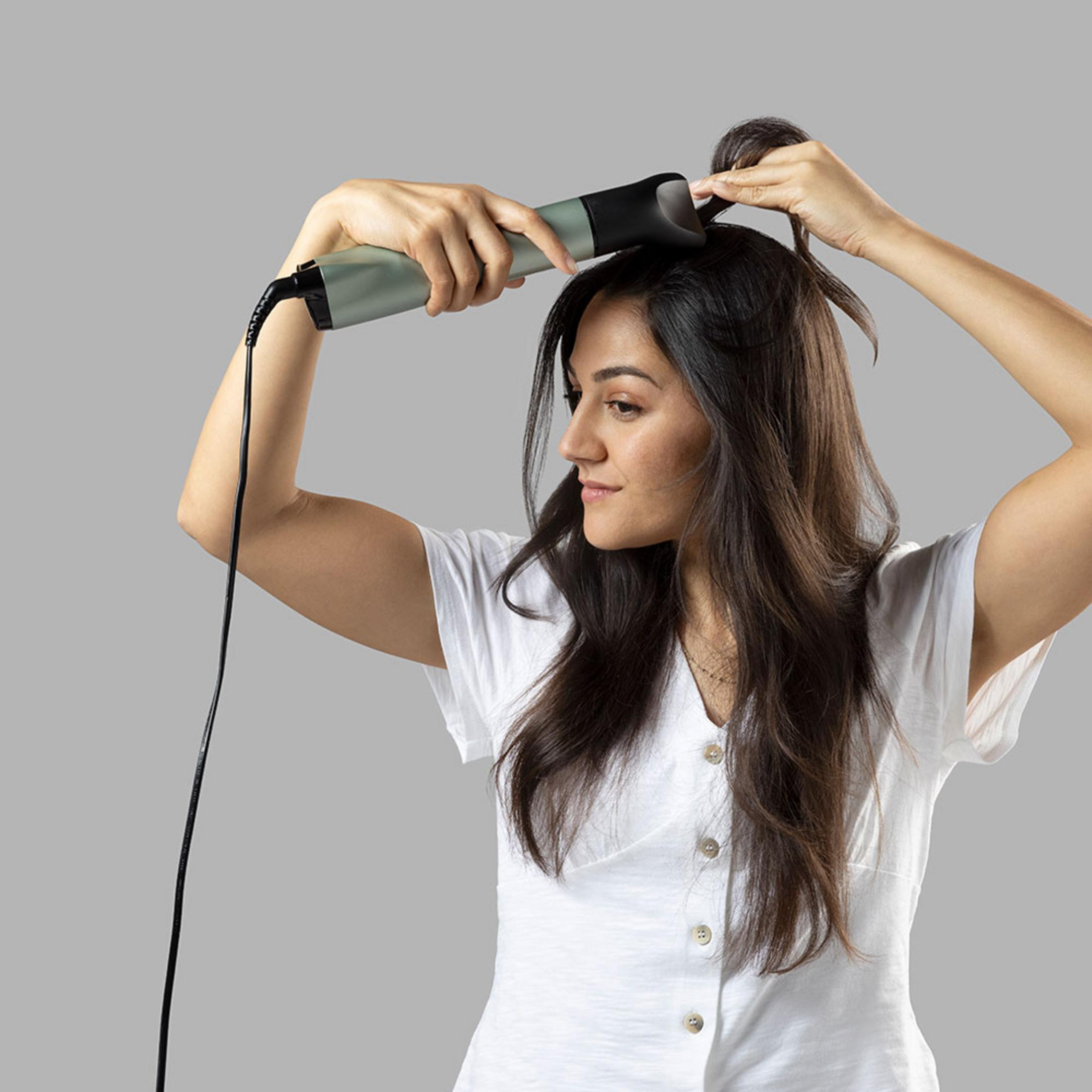 Kobieta z długimi ciemnymi włosami stylizuje włosy prostownicą.