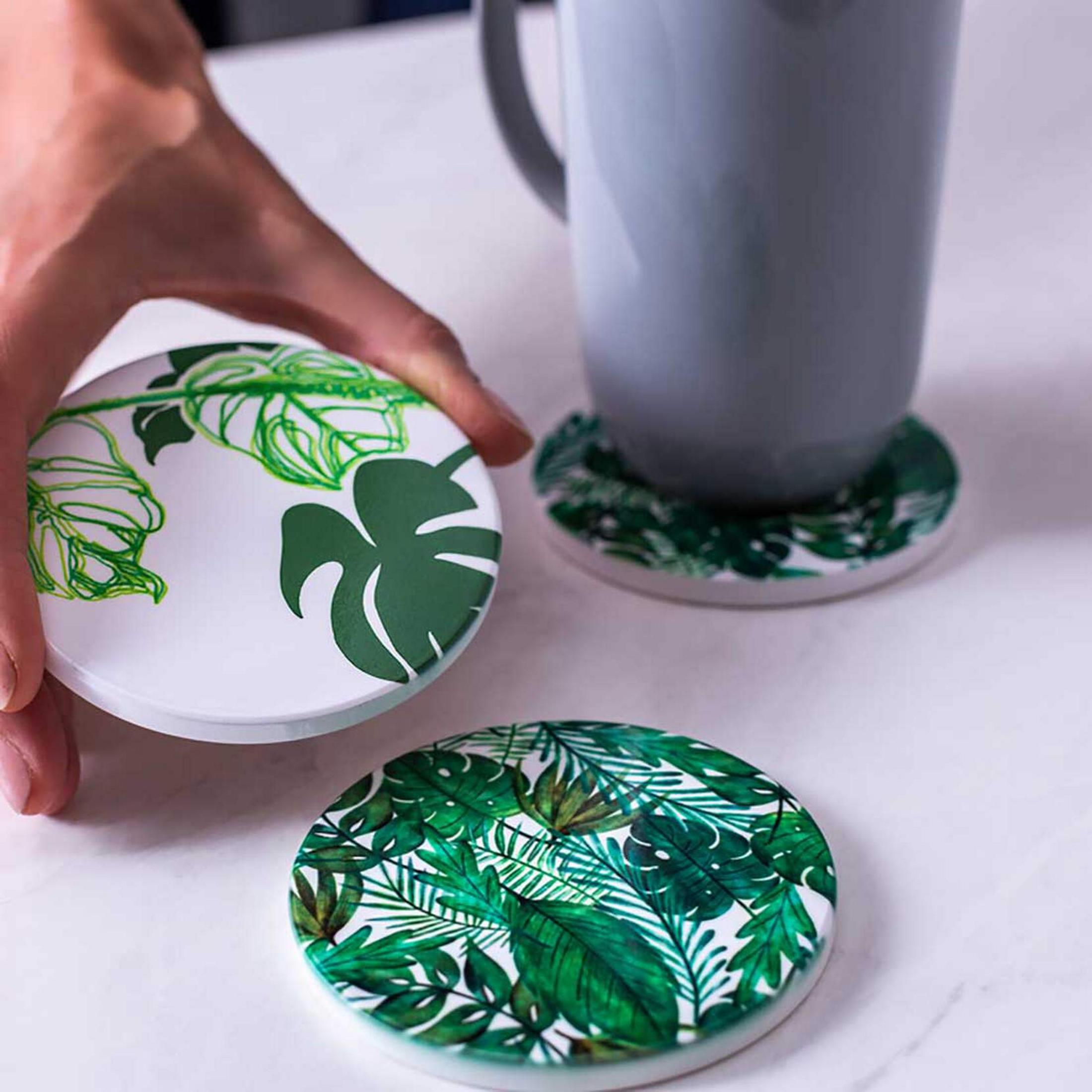 Hand hält Untersetzer mit grünen Blättern, neben Tasse auf Marmortisch.