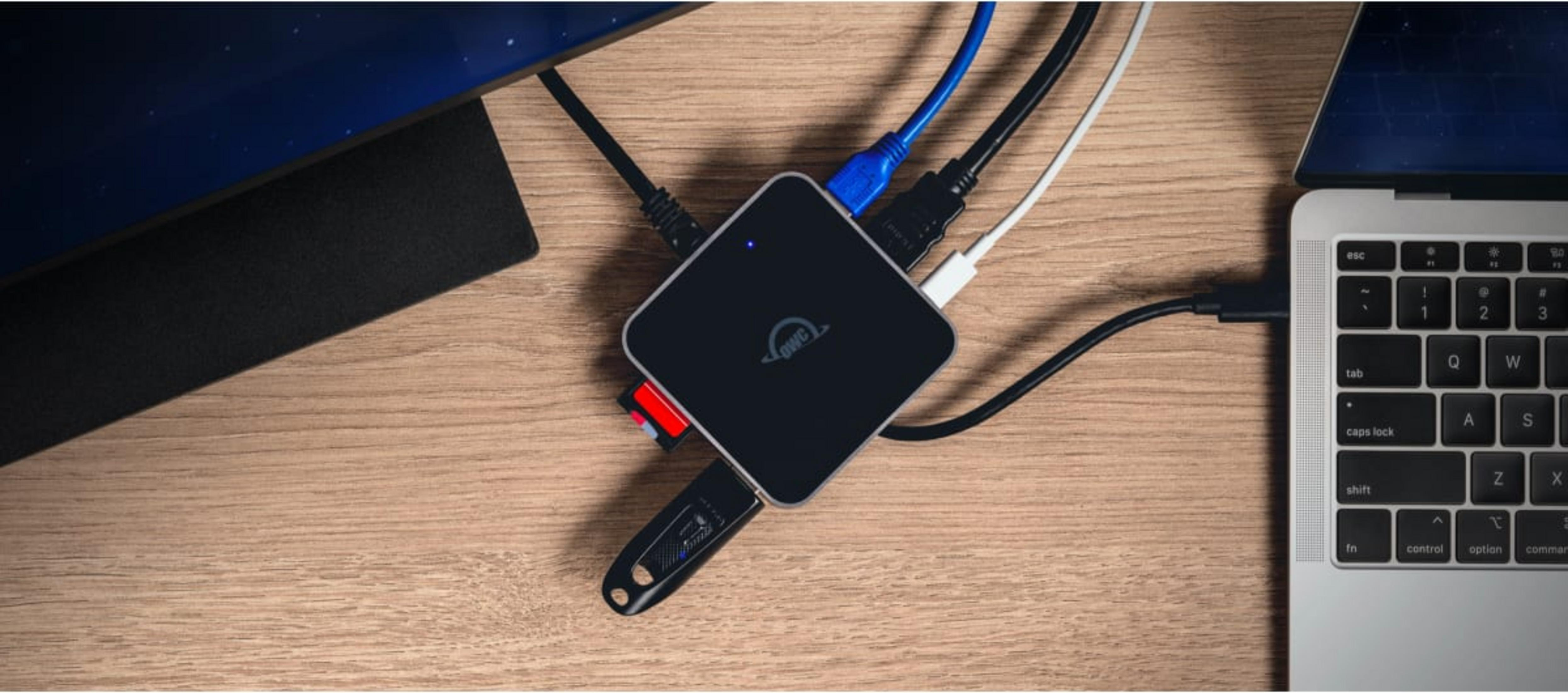 Een zwarte OWC-hub verbonden met kabels en een USB-stick op een houten bureau.