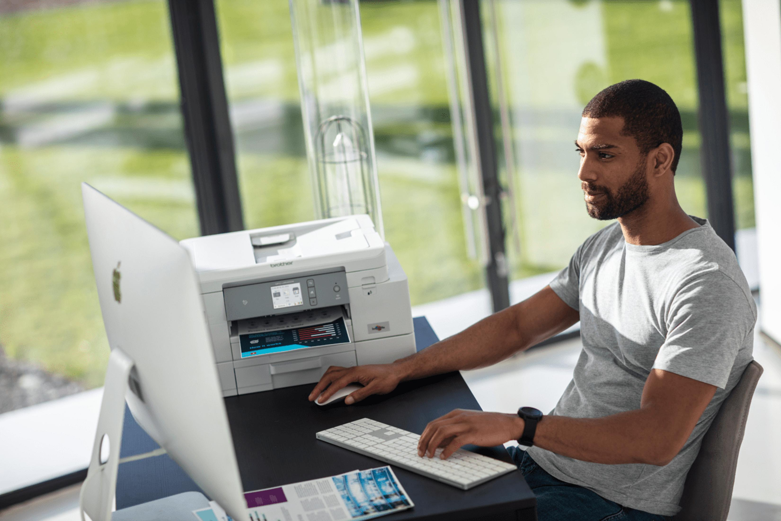 Mężczyzna przy biurku z komputerem i drukarką, zielone drzewa na zewnątrz.