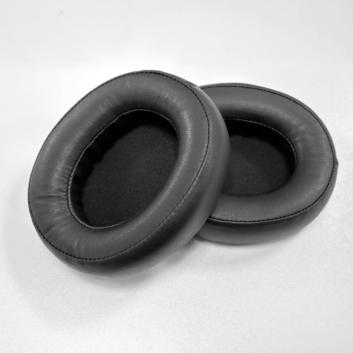 Dwie czarne, owalne nauszniki ze skóry ekologicznej leżą obok siebie na białej powierzchni.