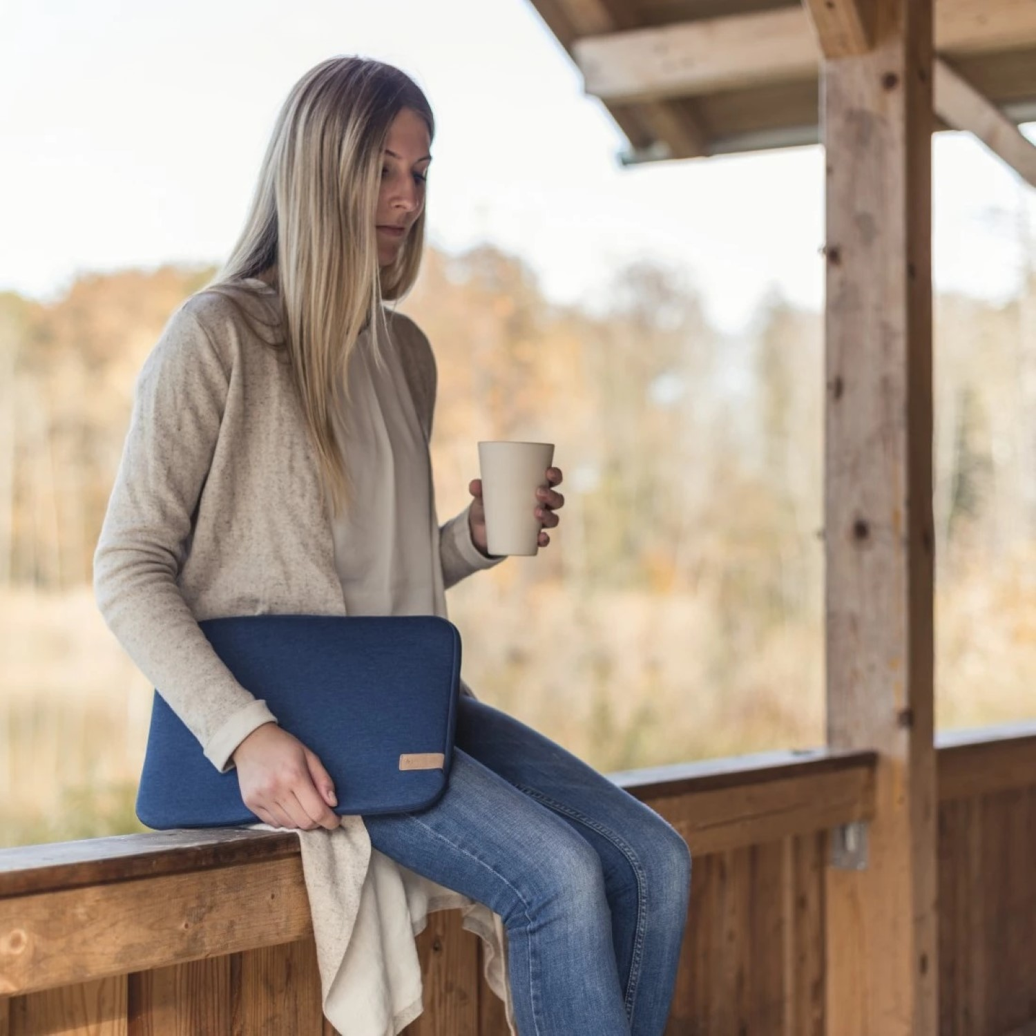 Frau sitzt auf Holzgeländer mit Laptoptasche und Becher, Herbstlandschaft.