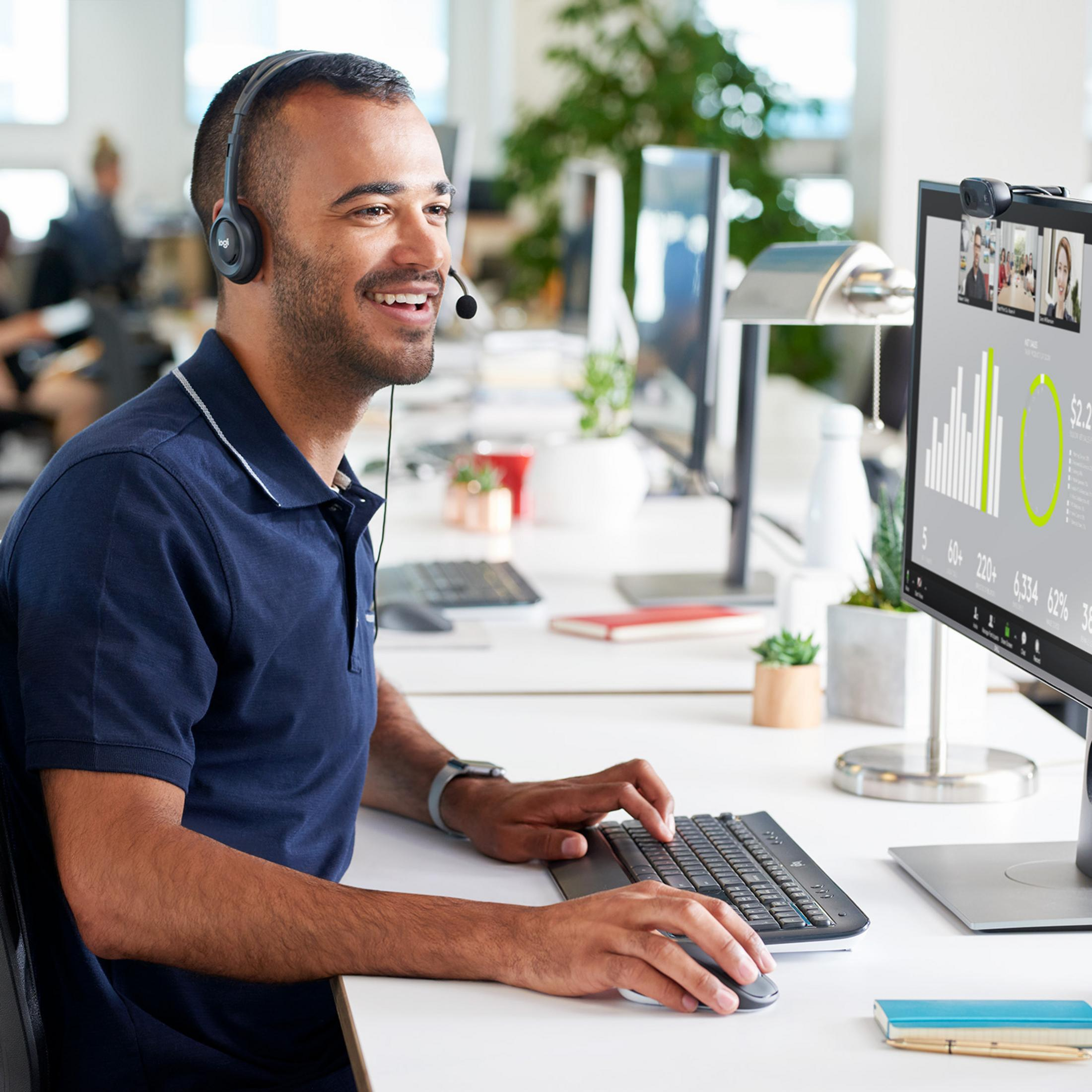 Mann mit Headset im Büro lächelt, tippt auf Tastatur, schaut auf Monitor mit Diagrammen.